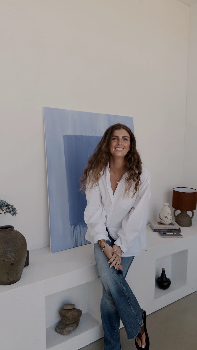 A woman with long wavy brown hair smiling while sitting on a white shelf in a bright room, wearing a loose white shirt and blue jeans, with decorative vases and books behind her, and a large blue abstract painting on the wall.