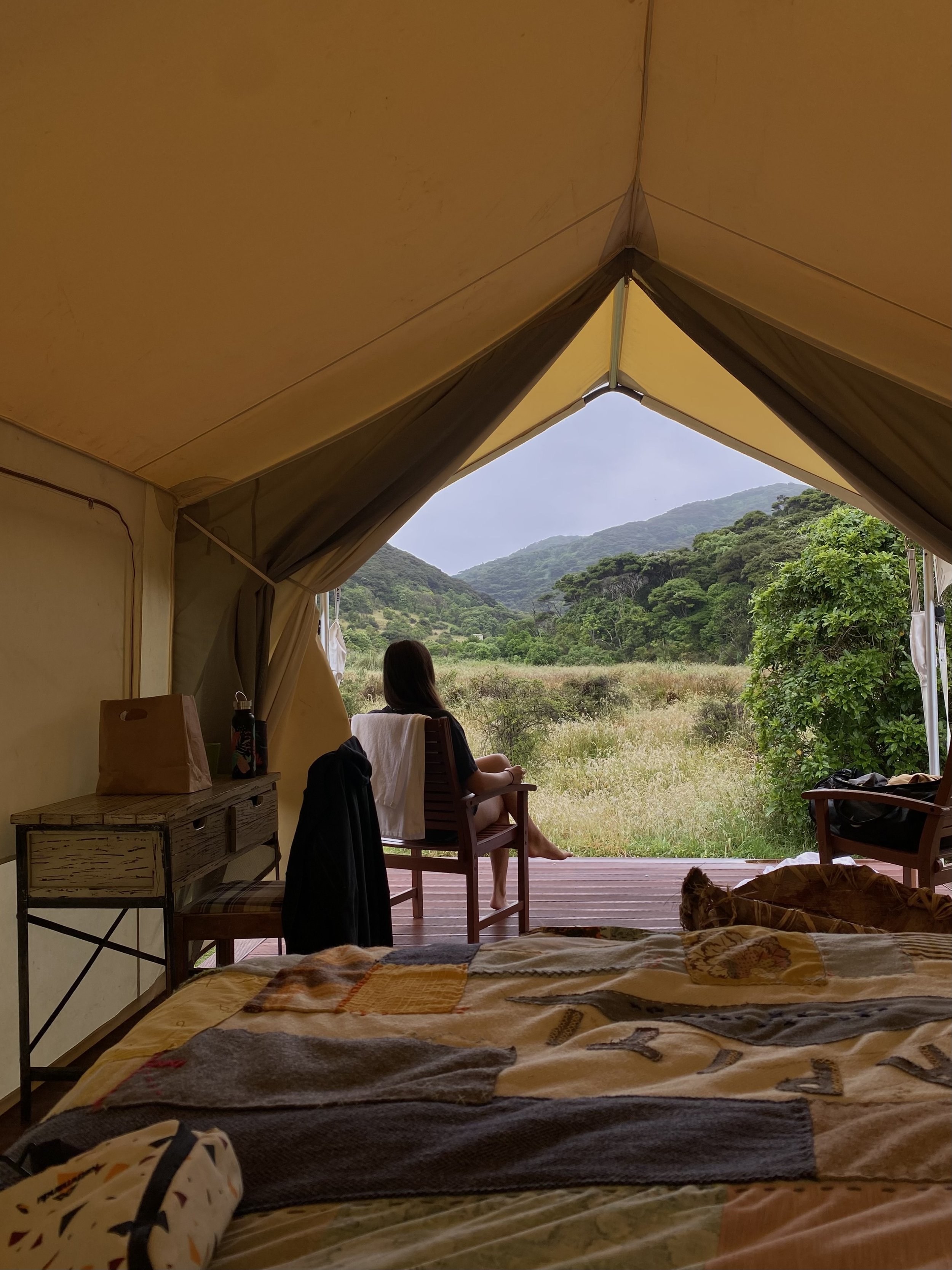 Inside a tent showing a view of green hills and trees through an open flap, with two women sitting on chairs outside, and a bed with patterned blankets inside.