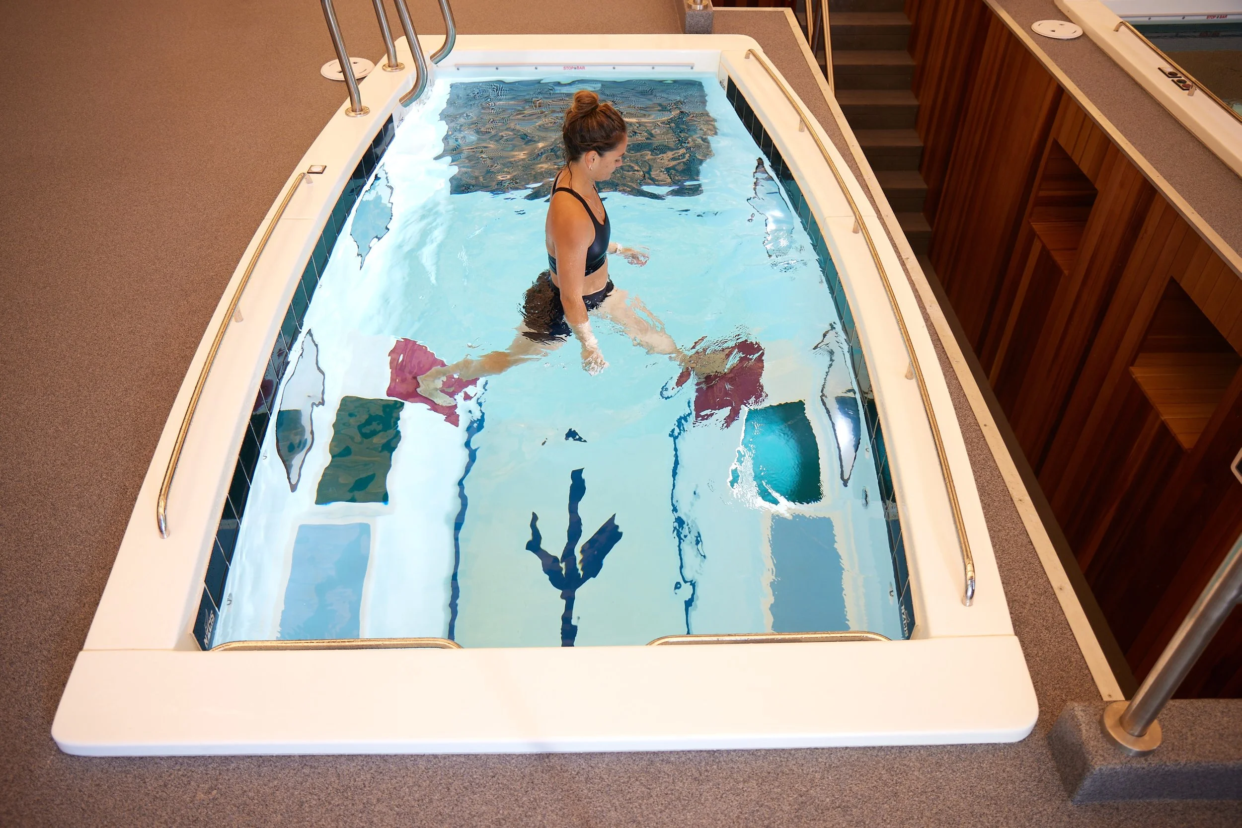 A woman in a black swimsuit standing in a small indoor pool with colorful tiled walls, surrounded by a beige carpeted floor and wooden paneling.