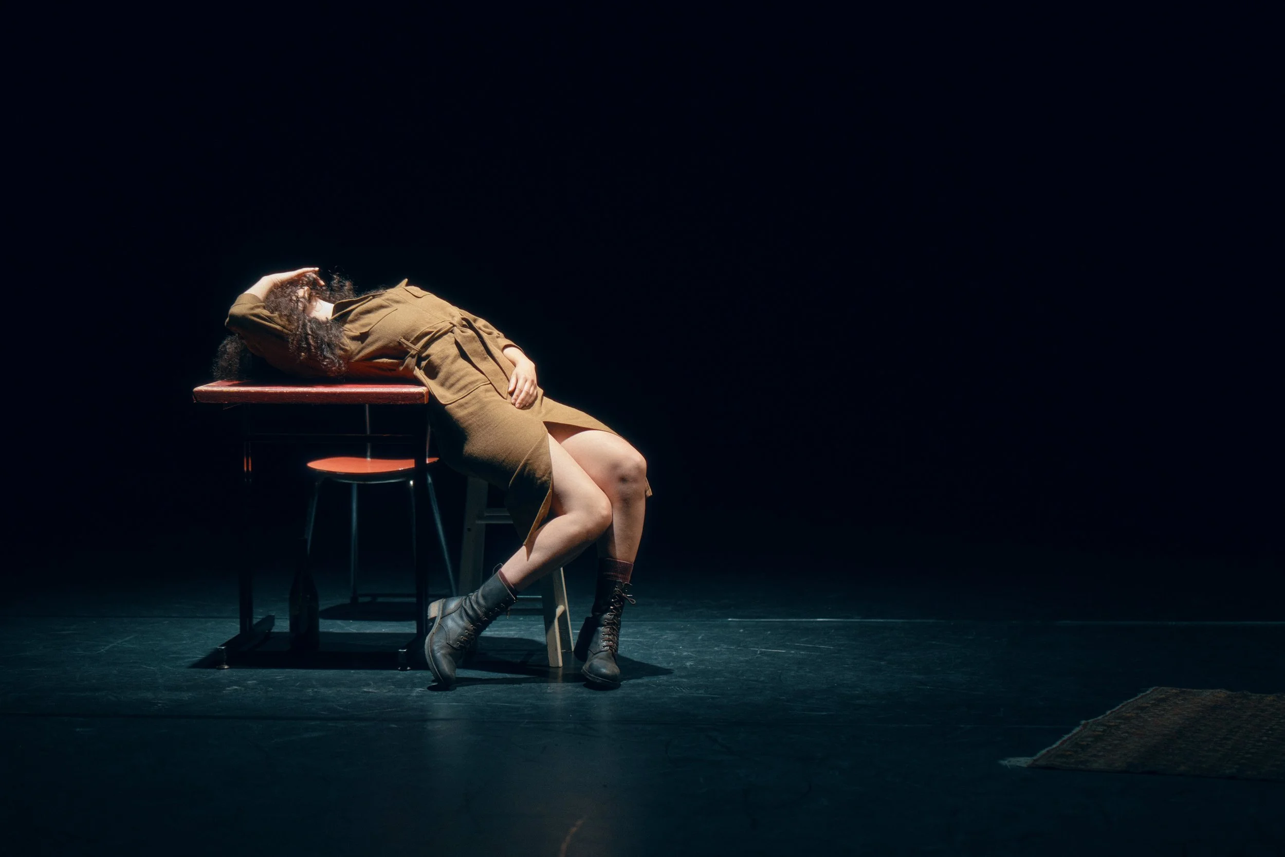 Une femme en vêtements marron, assise sur une chaise, penchée en arrière avec la tête reposant sur une table, dans un éclairage sombre.