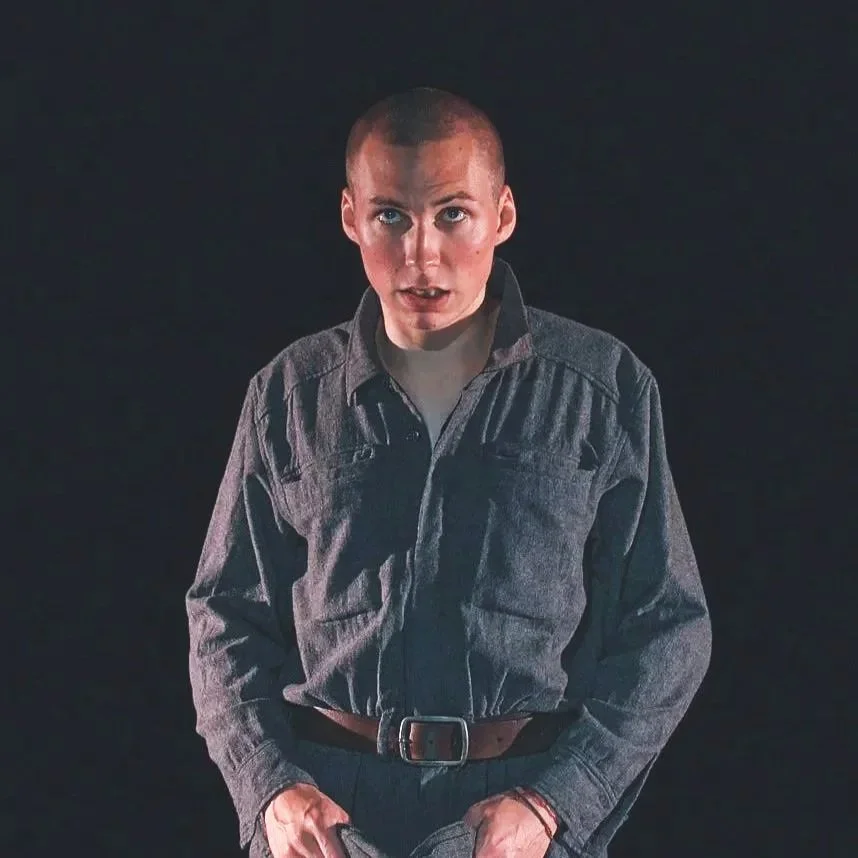 Jeune homme avec tête rasée, portant une chemise en denim gris, devant un fond noir, regardant l'objectif.