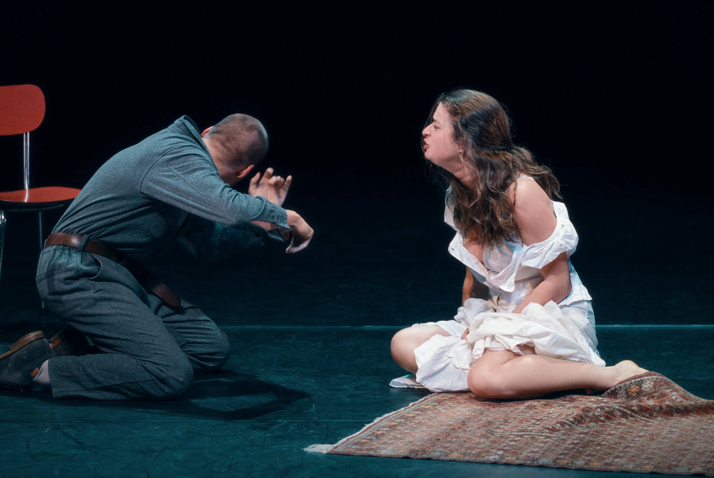 Une scène de théâtre avec un homme à genoux qui semble plaider ou implorer et une femme assise sur un tapis qui semble réagir émotionnellement.