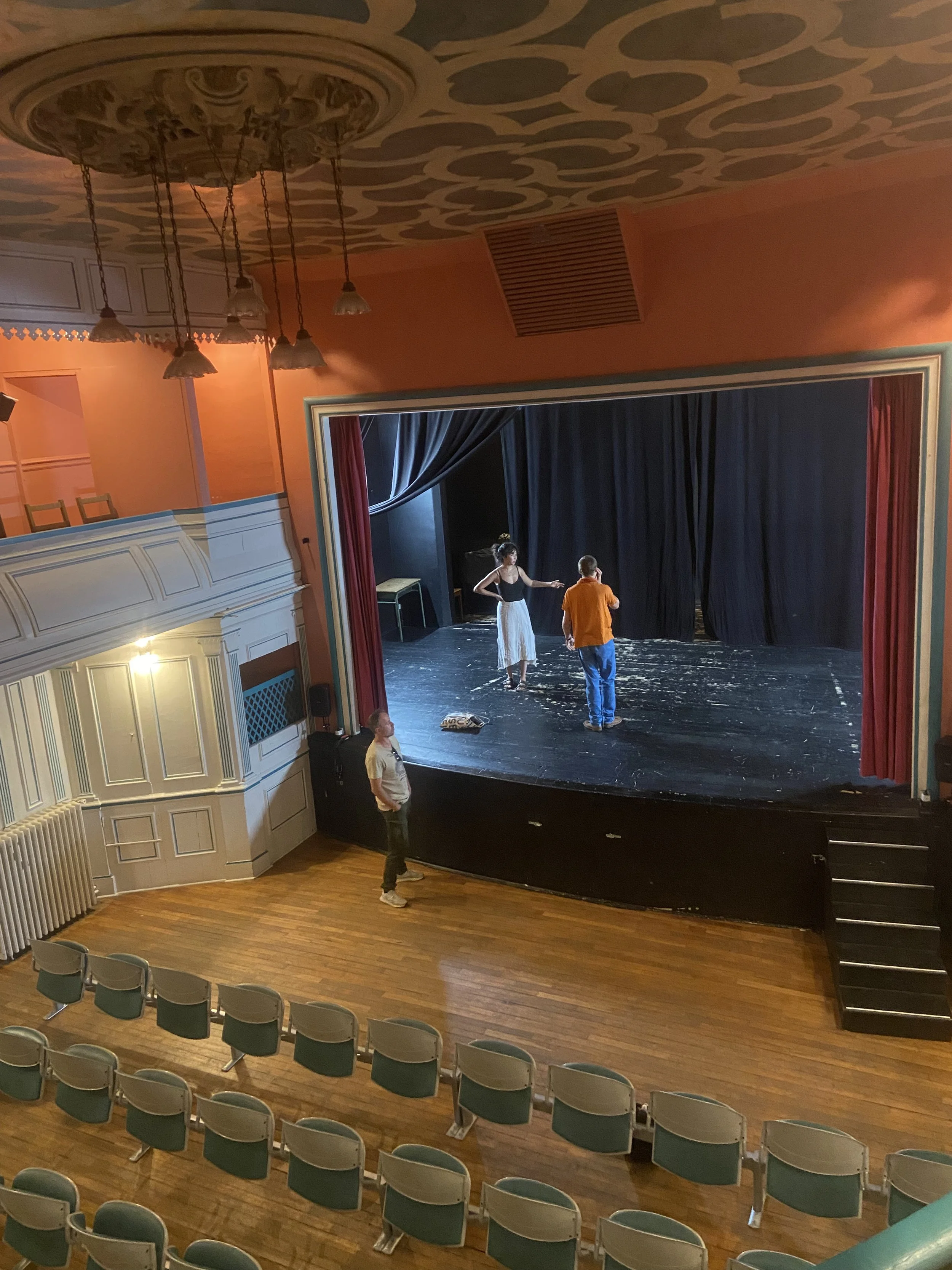 Une scène de théâtre avec trois personnes sur la scène, deux en train de discuter et une observant depuis le public. Le théâtre est vide avec des sièges bleus et des lumières tamisées.