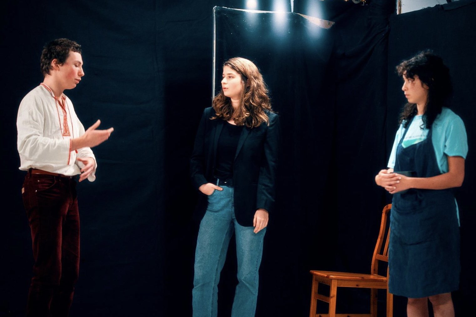 Trois personnes discutant dans une pièce sombre, deux femmes et un homme, avec une chaise en bois à l'arrière-plan.