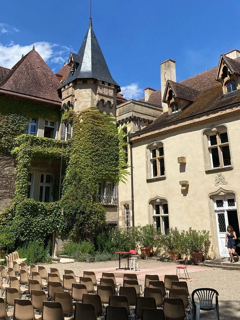 Un bâtiment historique avec une tour, des fenêtres anciennes, et beaucoup de végétation grimpante, avec une zone en plein air équipée de chaises et d'une table, un ciel bleu avec quelques nuages.