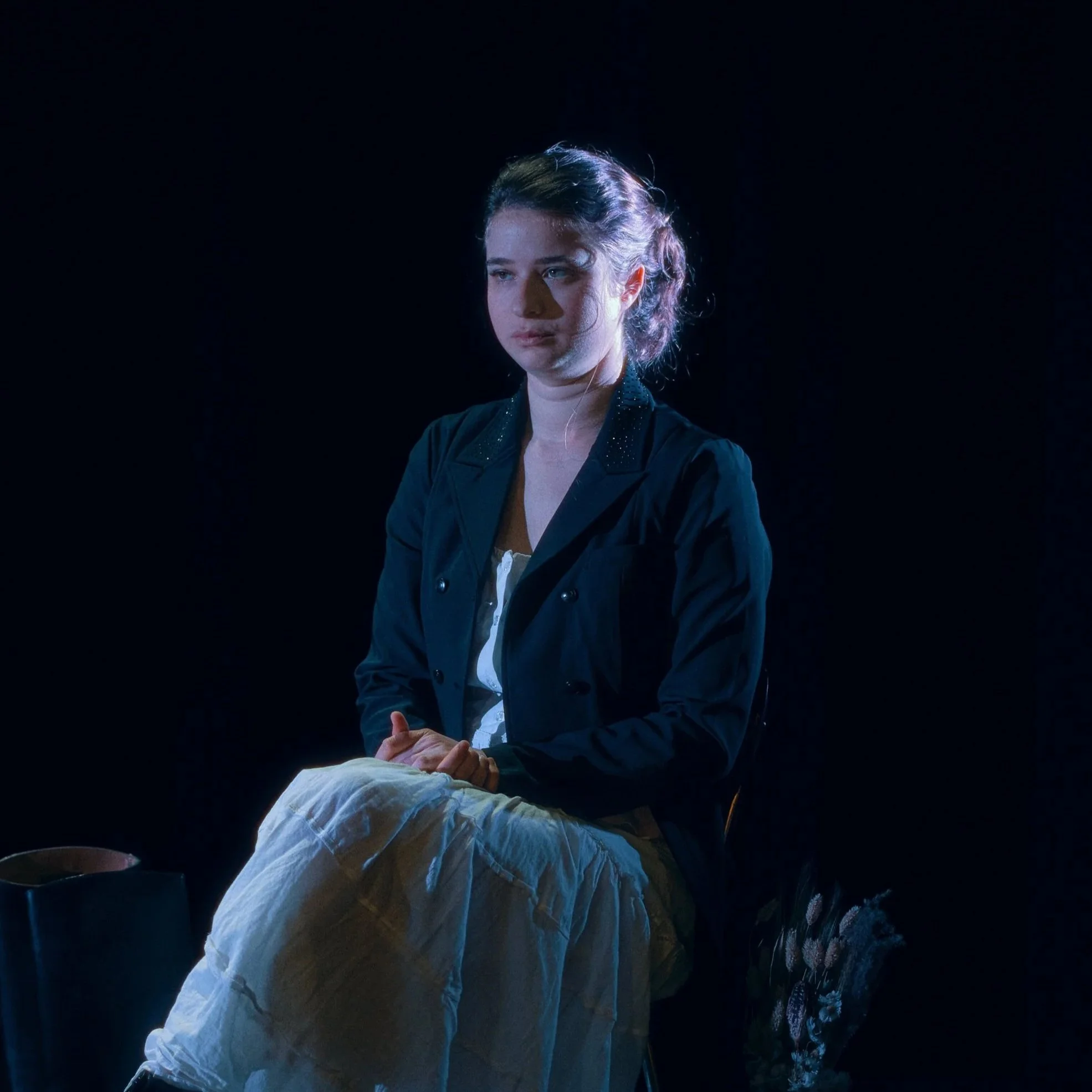 Femme assise dans une lumière tamisée, portant un manteau noir et une jupe claire, avec un sac noir à côté, dans un contexte sombre.