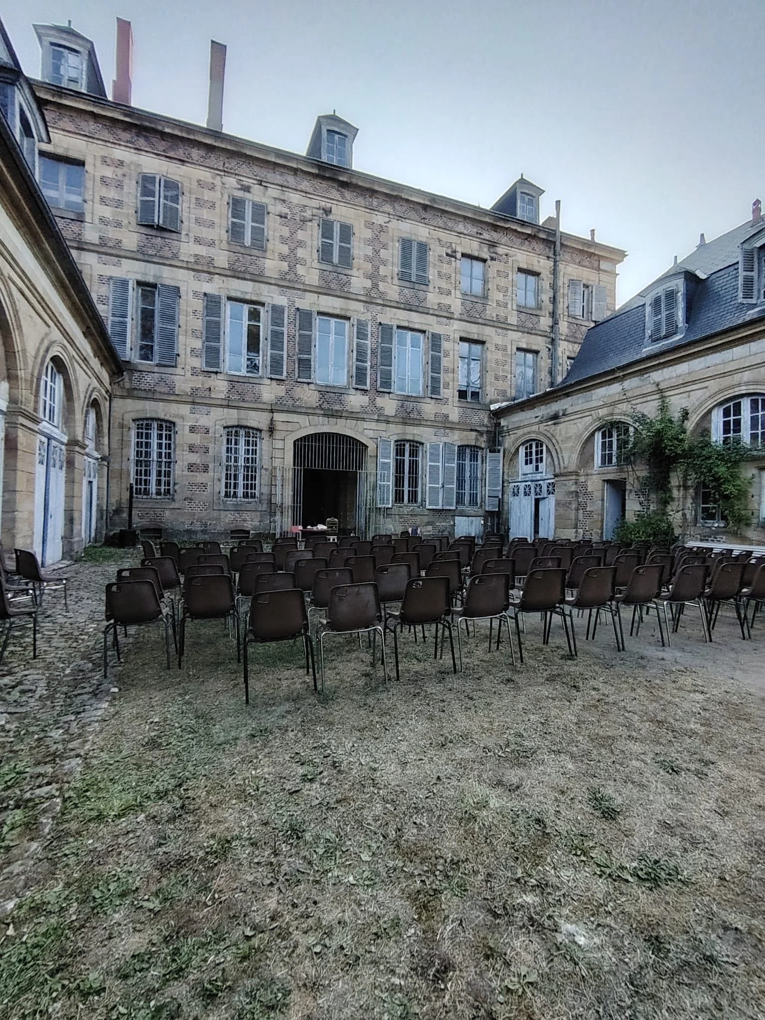 Un cour d'école ou de cour intérieure avec plusieurs chaises alignées sur le sol en terre meuble, entourée de bâtiments en pierre avec fenêtres à volets, sous un ciel clair.