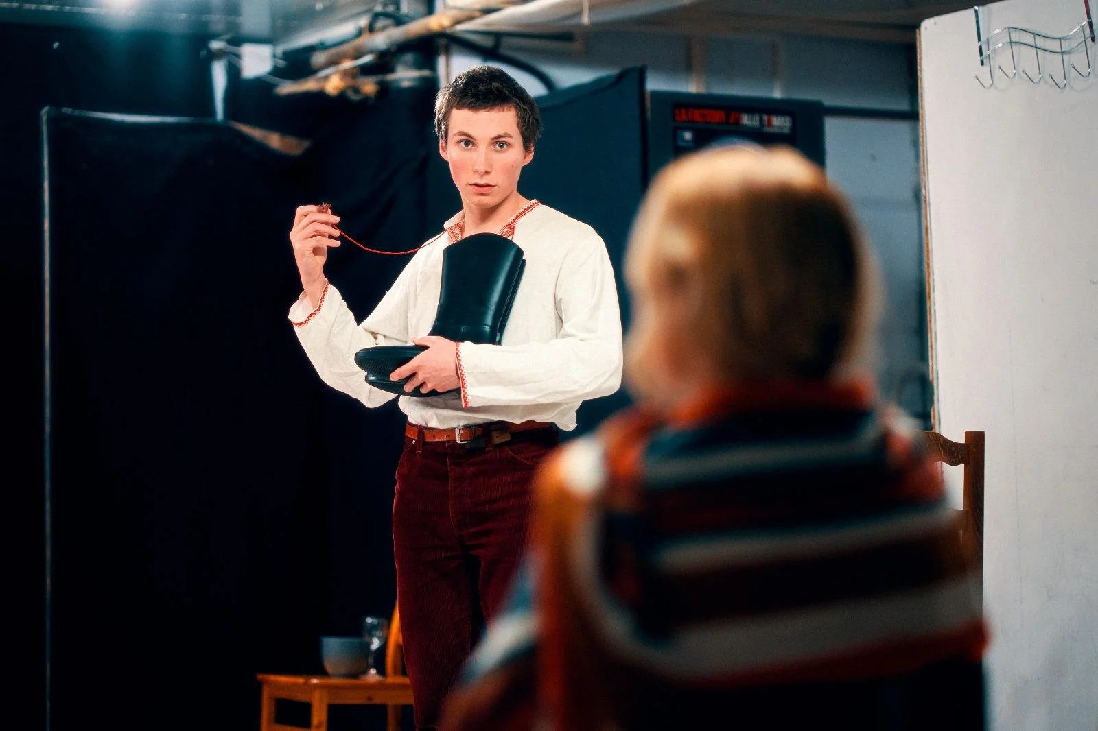 Un jeune homme avec des cheveux courts et une chemise blanche tient une botte en caoutchouc et un fil électrique dans une pièce. Il regarde vers l'avant avec une expression sérieuse, un adulte est assis devant lui, vu de dos, portant un vêtement à rayures.