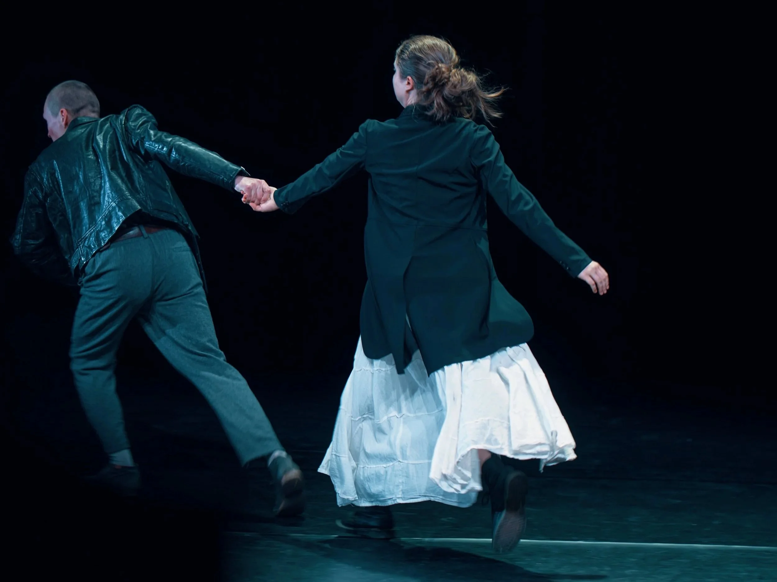Deux personnes dansent sur scène, tenant la main dans une posture dynamique contre un fond noir.