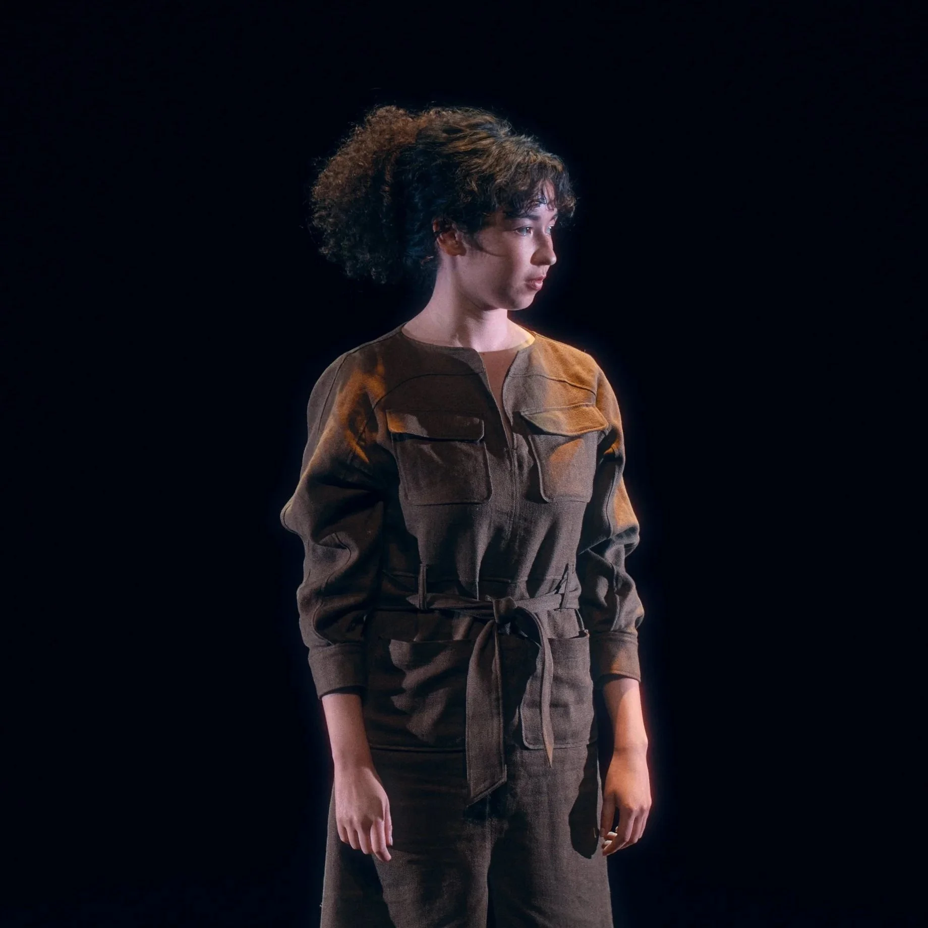 Jeune femme avec des cheveux bouclés, portant un uniforme militaire marron, se tenant contre un fond noir.