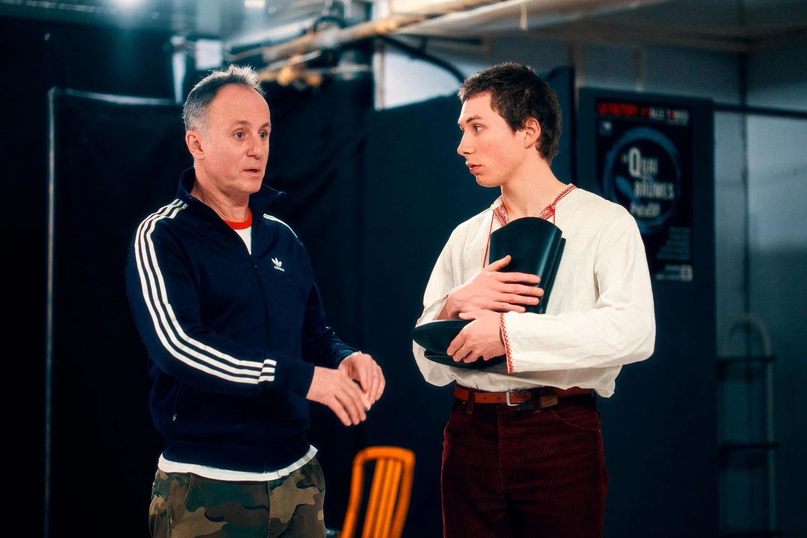 Deux hommes en conversation dans un endroit intérieur, l'un porte une veste adidas et l'autre une chemise blanche avec des détails rouges, tenant un étui ou un objet noir.