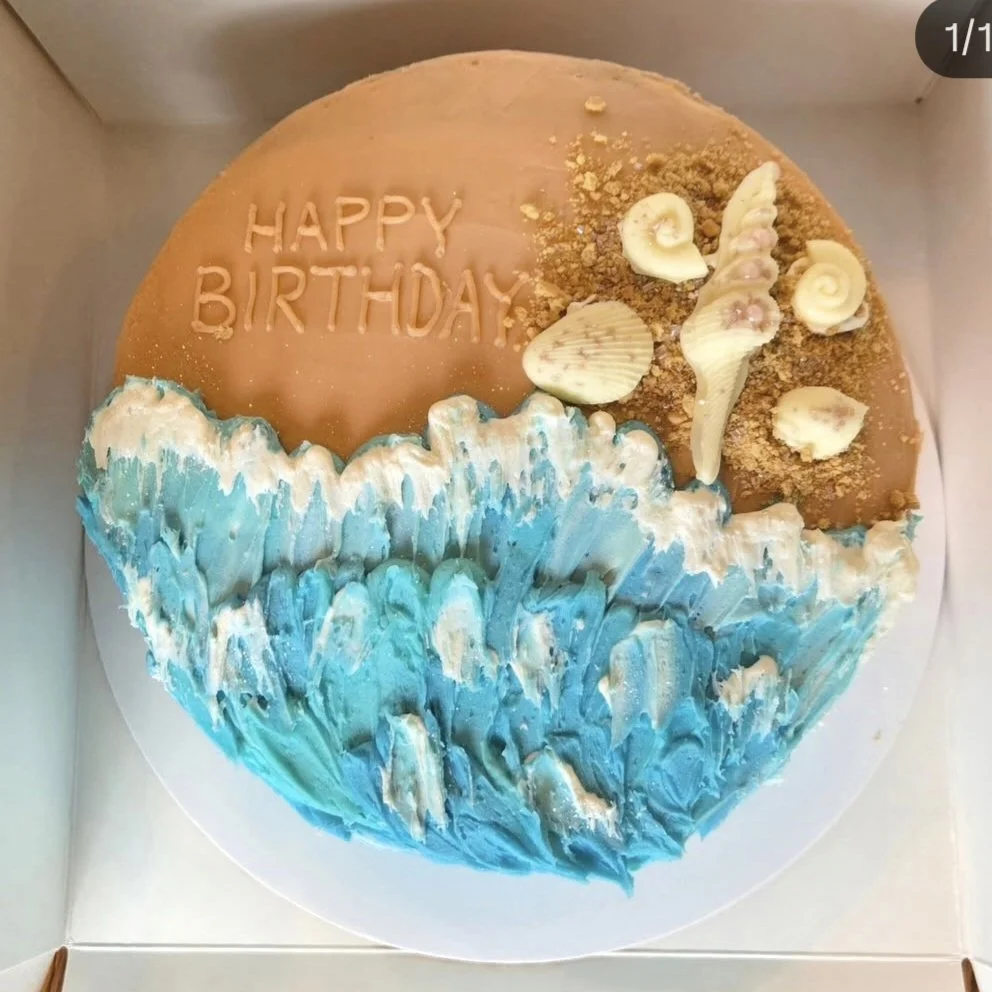 Round birthday cake decorated with ocean waves made of blue and white frosting at the bottom, topped with shells, sand, and caramel-colored icing with the words "HAPPY BIRTHDAY" written on it.
