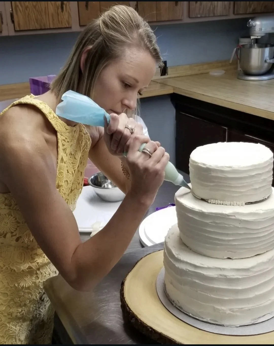 A woman wearing a yellow lace dress decorates a three-tiered white wedding cake with a piping bag in a kitchen.