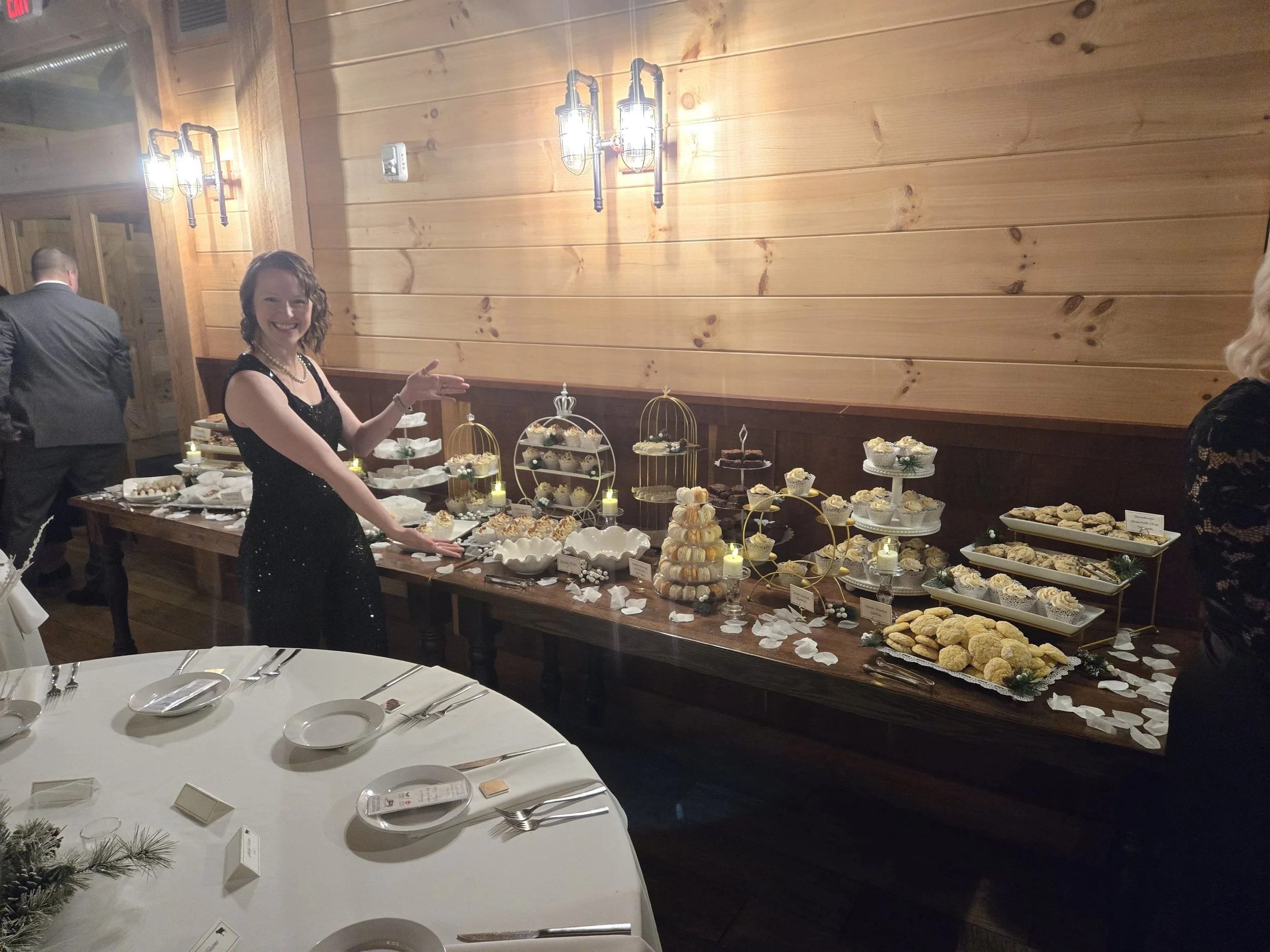 Woman in black dress pointing at a large dessert table with assorted cookies and cupcakes at a holiday or wedding reception.