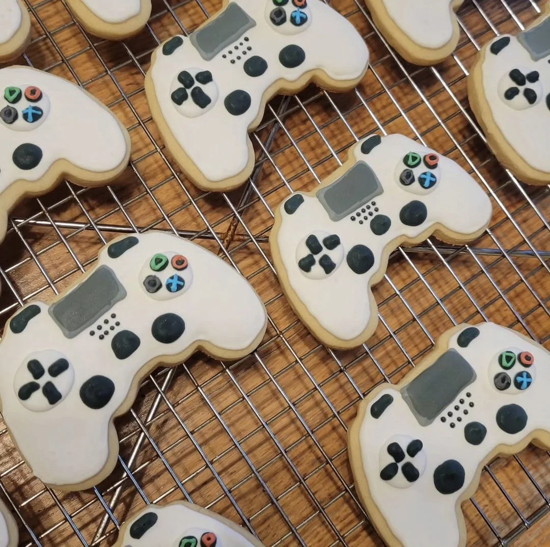 Cookies decorated as video game controllers with white icing, colorful buttons, and gray sticks, on a wire cooling rack.