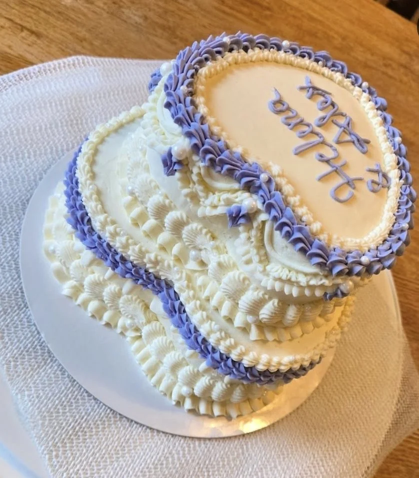 A tiered, heart-shaped birthday cake decorated with white and light purple frosting, featuring piped decorations and pearls, with 'Happy Birthday' written on top.