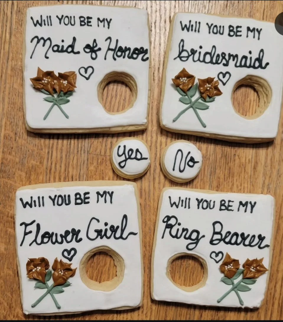 Cookies decorated with white icing, each with a different wedding-related proposal and floral decorations: "Will you be my maid of honor," "Will you be my bridesmaid," "Will you be my flower girl," and "Will you be my ring bearer."