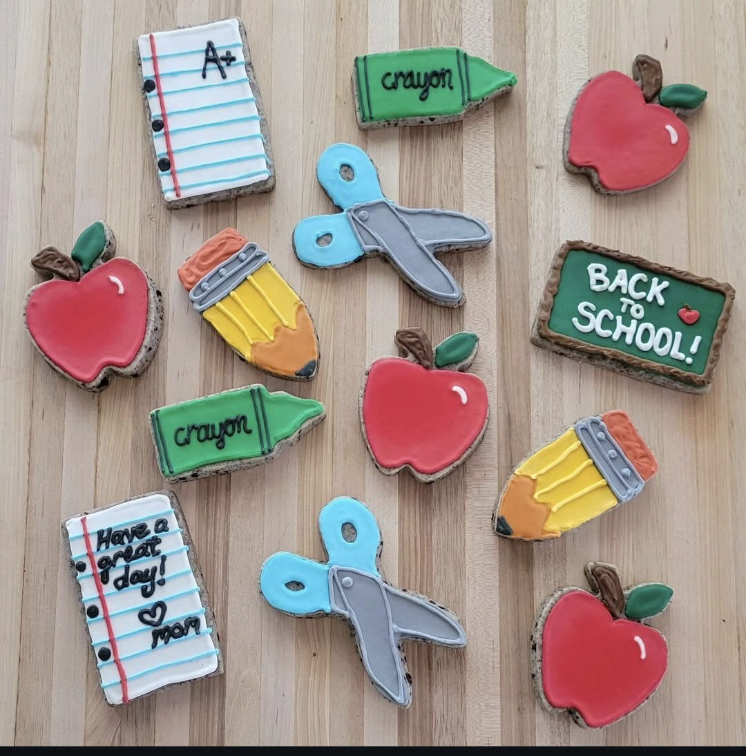 Decorated cookies resembling school supplies and messages for back to school, including red apples, yellow pencils, green crayons, blue and gray butterflies, a chalkboard with "Back to School!", and notebooks with messages.