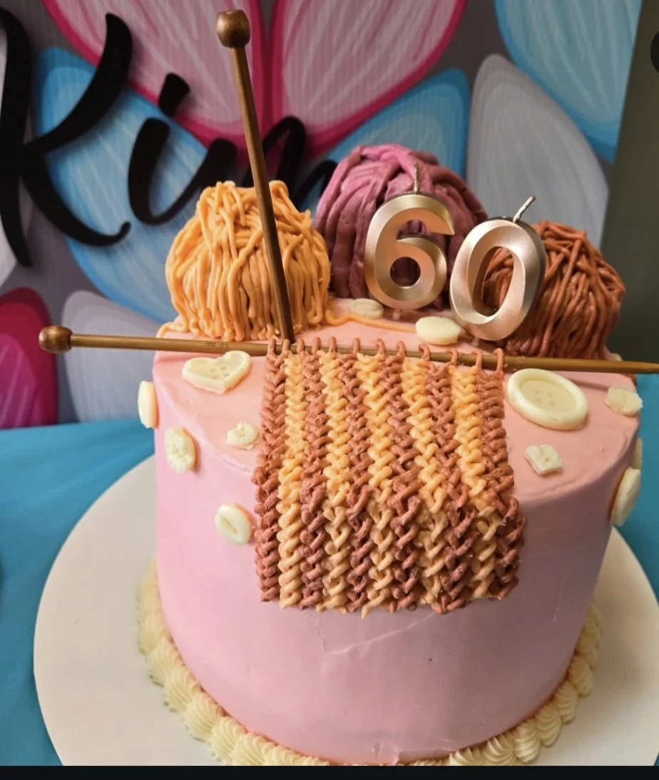 A pink birthday cake decorated with found yarn balls, a knitting needle with a golden tip, and a pocket of yarn hanging off the side. It has the number 60 as a cake topper and some small white decorative elements. Background has colorful hearts and the word 'love'.