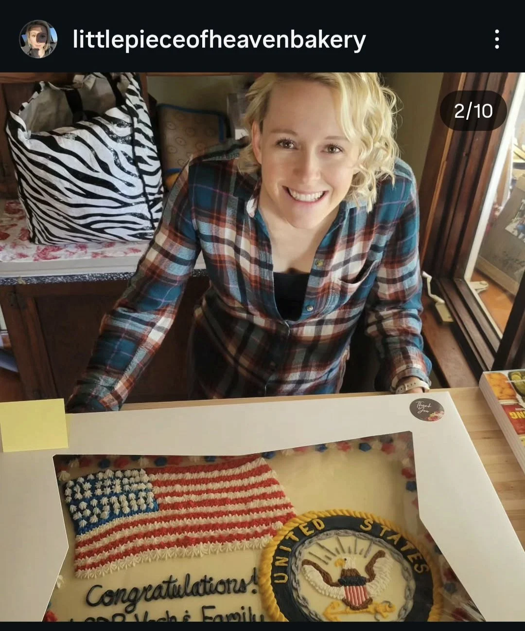 A woman with short curly blonde hair smiling, wearing a plaid shirt, standing behind a celebration cake featuring the United States Seal.