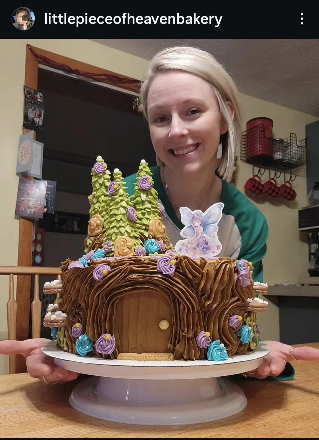A woman holding a whimsical, fairy-tale themed cake decorated with green trees, purple and blue flowers, and a fairy topper, in a home kitchen.