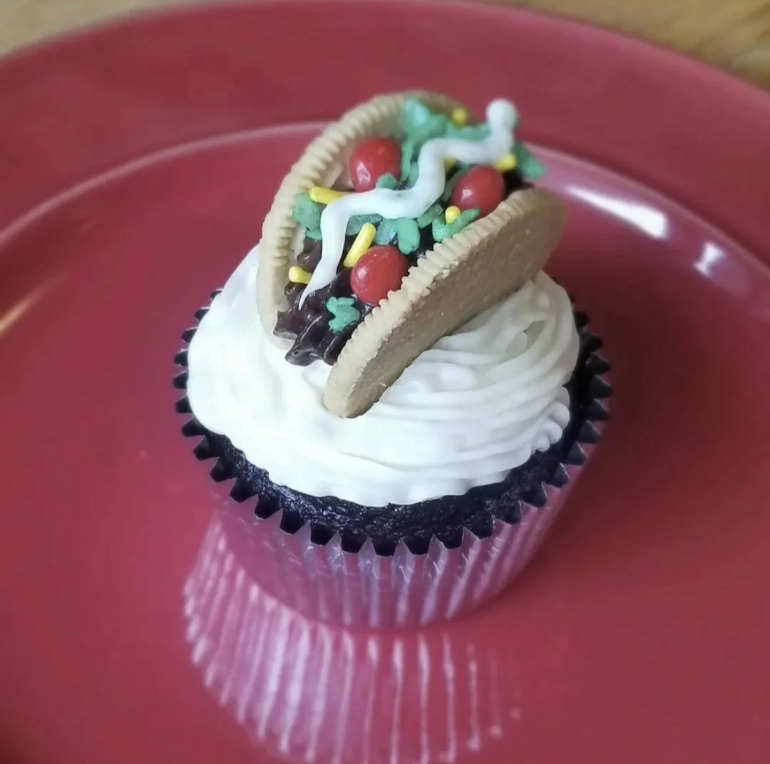 Cupcake with white frosting topped with a cookie, green sprinkles, red candies, and white and yellow icing decorations, on a red plate.