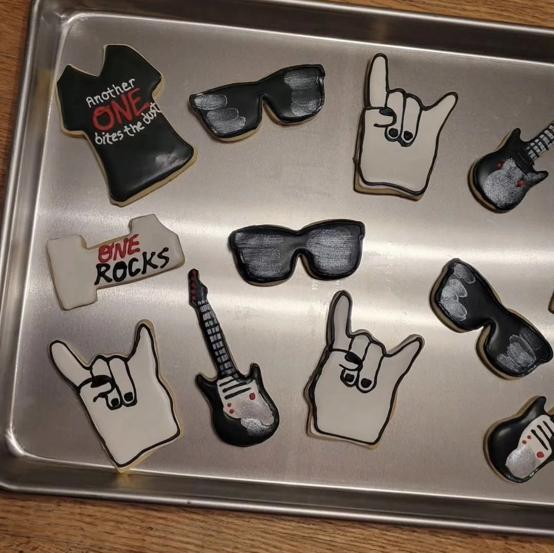 Assorted rock and roll-themed cookies with designs of guitars, sunglasses, hand signs, and text such as "Another One Bites the Dust" and "One Rocks" on a metal tray.