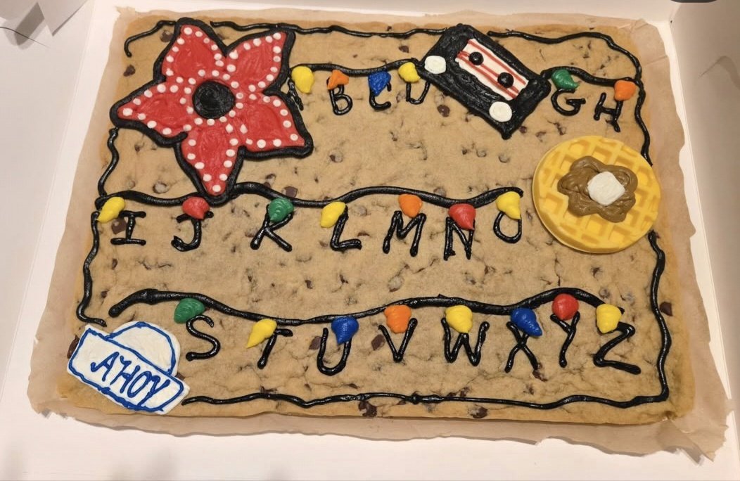A cake decorated to look like a vintage television set with a large red poinsettia flower, a bowl of waffle and ice cream, colorful string lights, and the alphabet written in black icing.