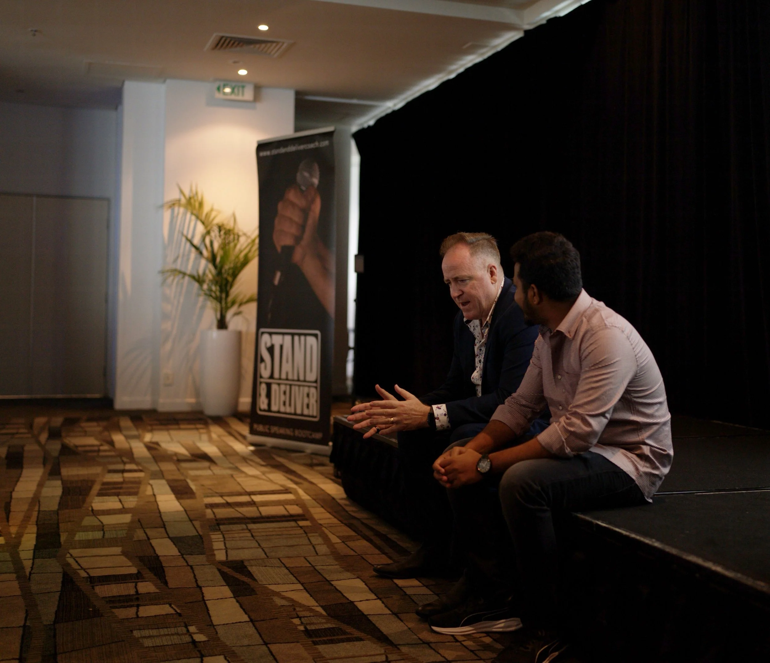 Two men sitting on the floor with their heads bowed in a dark conference room. There is a large potted plant and a vertical black banner with white text and an image of a person wearing a jacket in the background.
