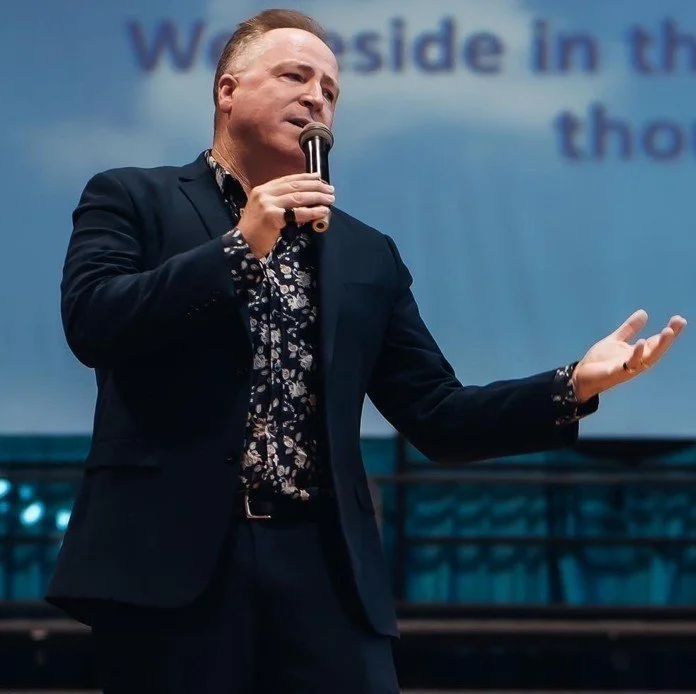 A man in a dark suit and floral shirt speaking into a microphone on stage with a projected presentation in the background.