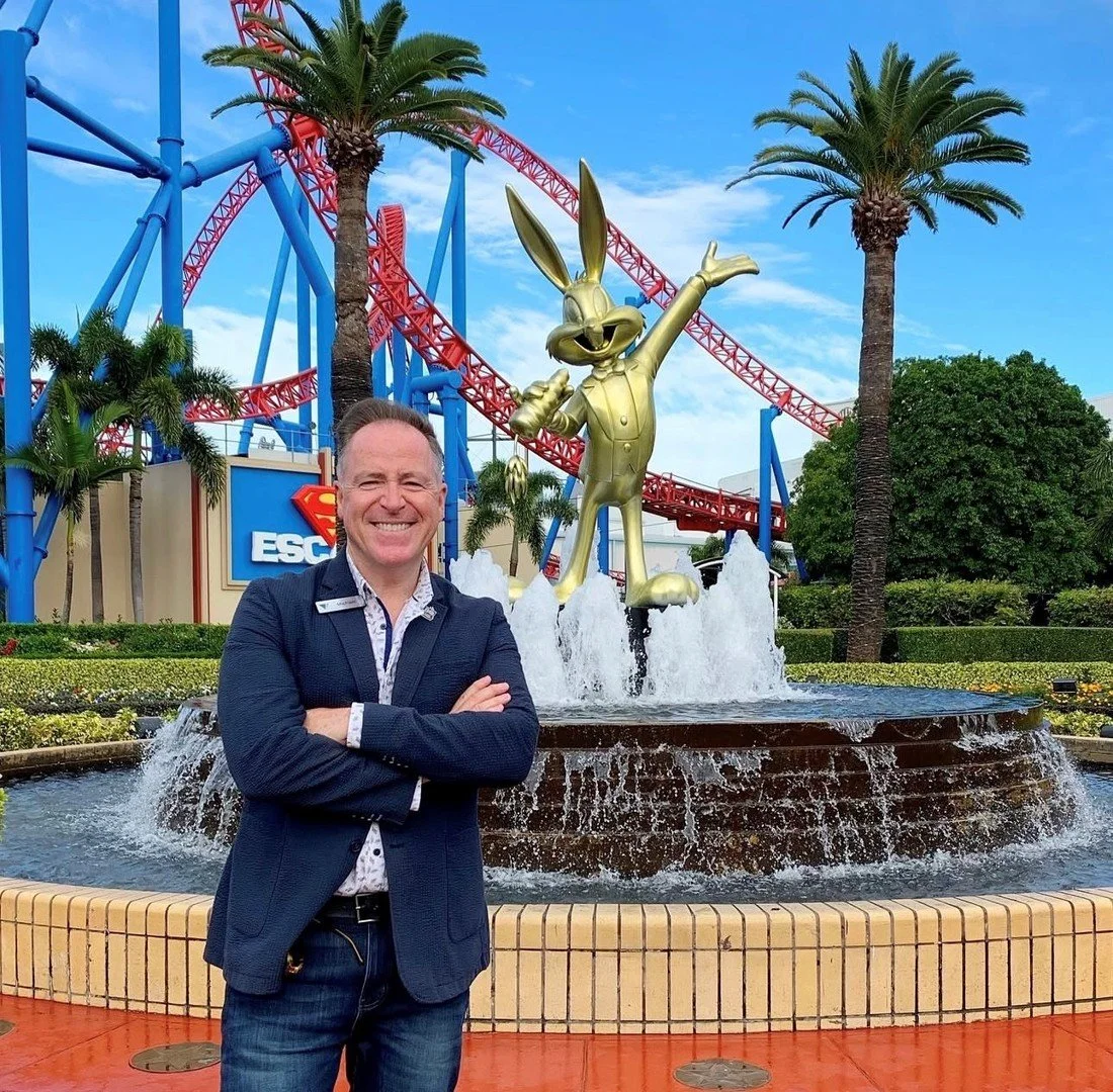 A man smiling with arms crossed in front of a Disneyland attraction featuring a large golden Bugs Bunny statue in a fountain with palm trees and roller coaster tracks in the background.