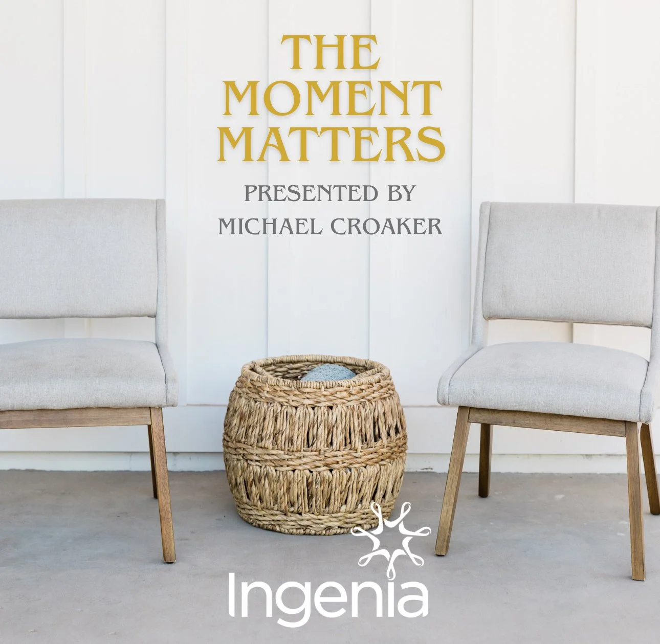 Event backdrop with the text 'The Moment Matters' presented by Michael Croaker, flanked by two light gray upholstered chairs and a woven ottoman in front. The setting appears to be a minimalist, modern indoor space.