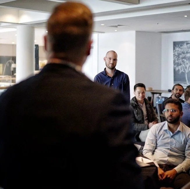 A man in a dark suit is speaking to a group of people in a modern indoor setting, with several men seated and listening attentively.