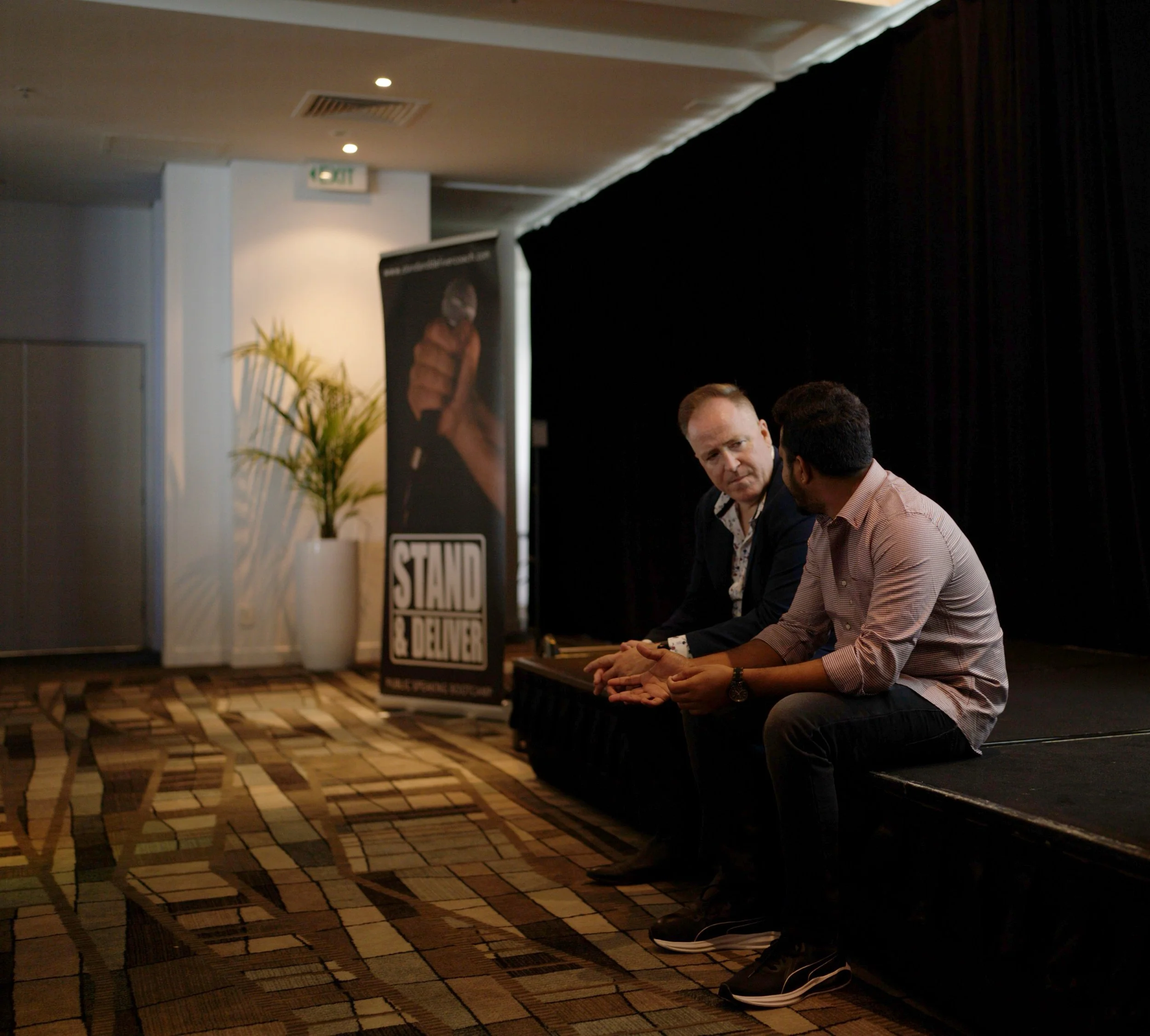 People sitting and talking at a table in a conference or event space, with a black curtain backdrop and a sign that says 'STAND' in the background.