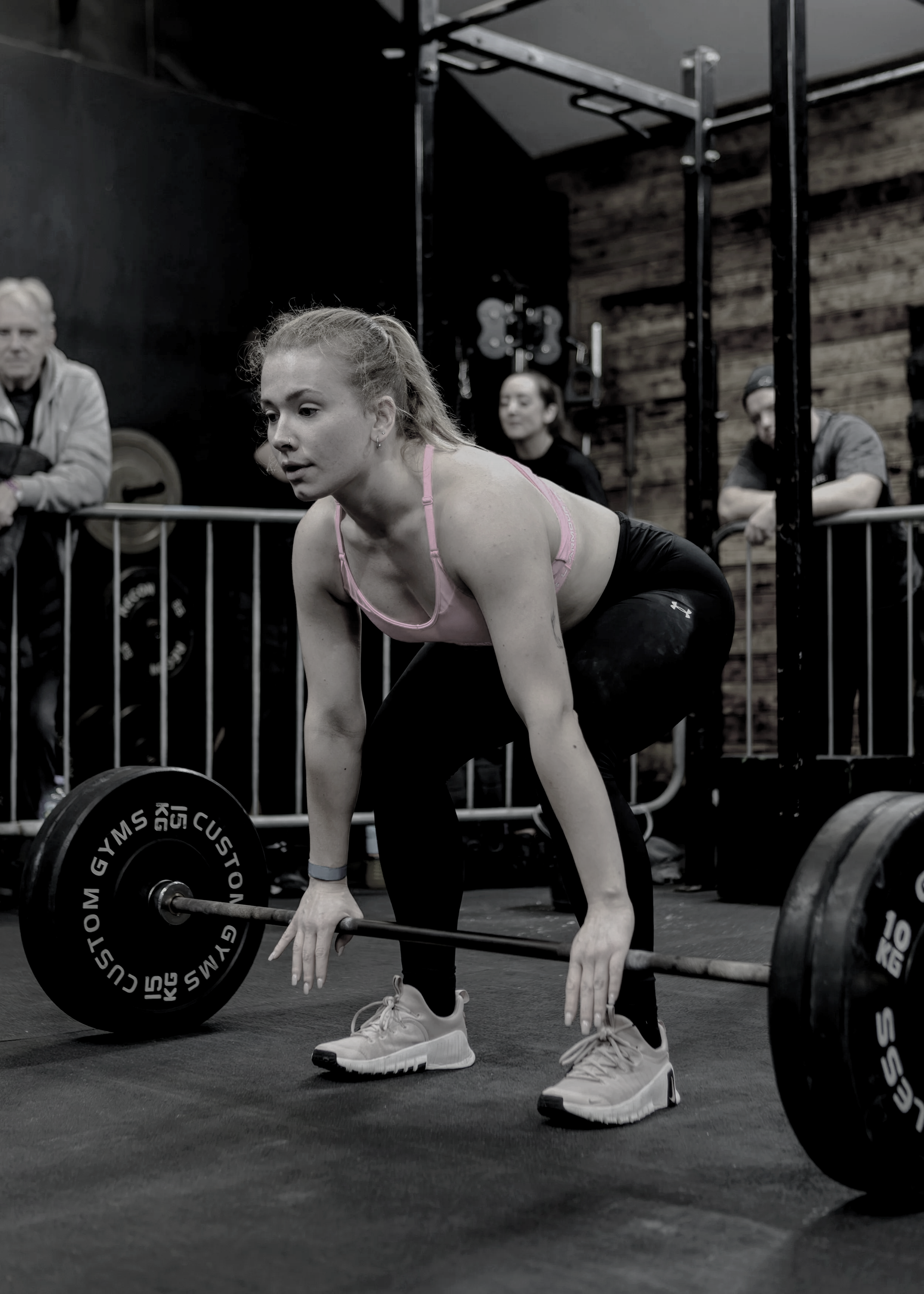 A woman in a pink sports bra and black leggings preparing to lift a barbell in a gym, with spectators watching.