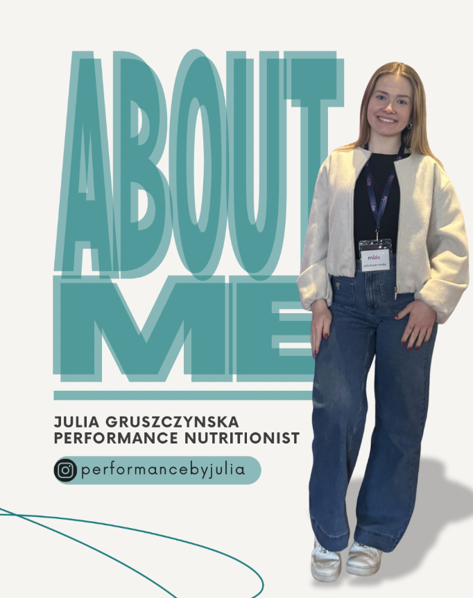 A woman standing next to a sign that says 'About Me'. She is smiling, wearing a light-colored jacket, dark shirt, jeans, and sneakers, with a name tag around her neck.