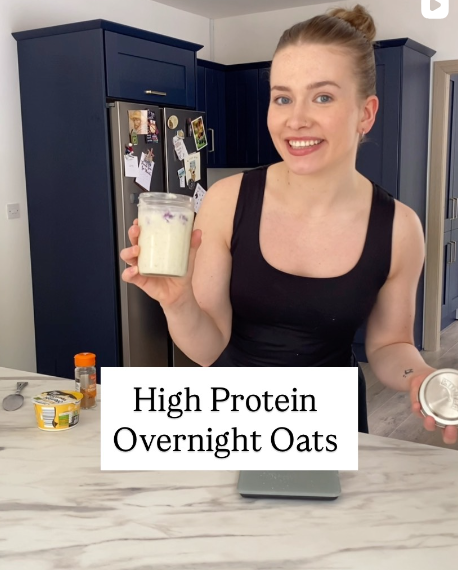 A woman in a black tank top holding a jar of overnight oats in one hand and a small container of ingredients in the other, standing in a kitchen with blue cabinets.
