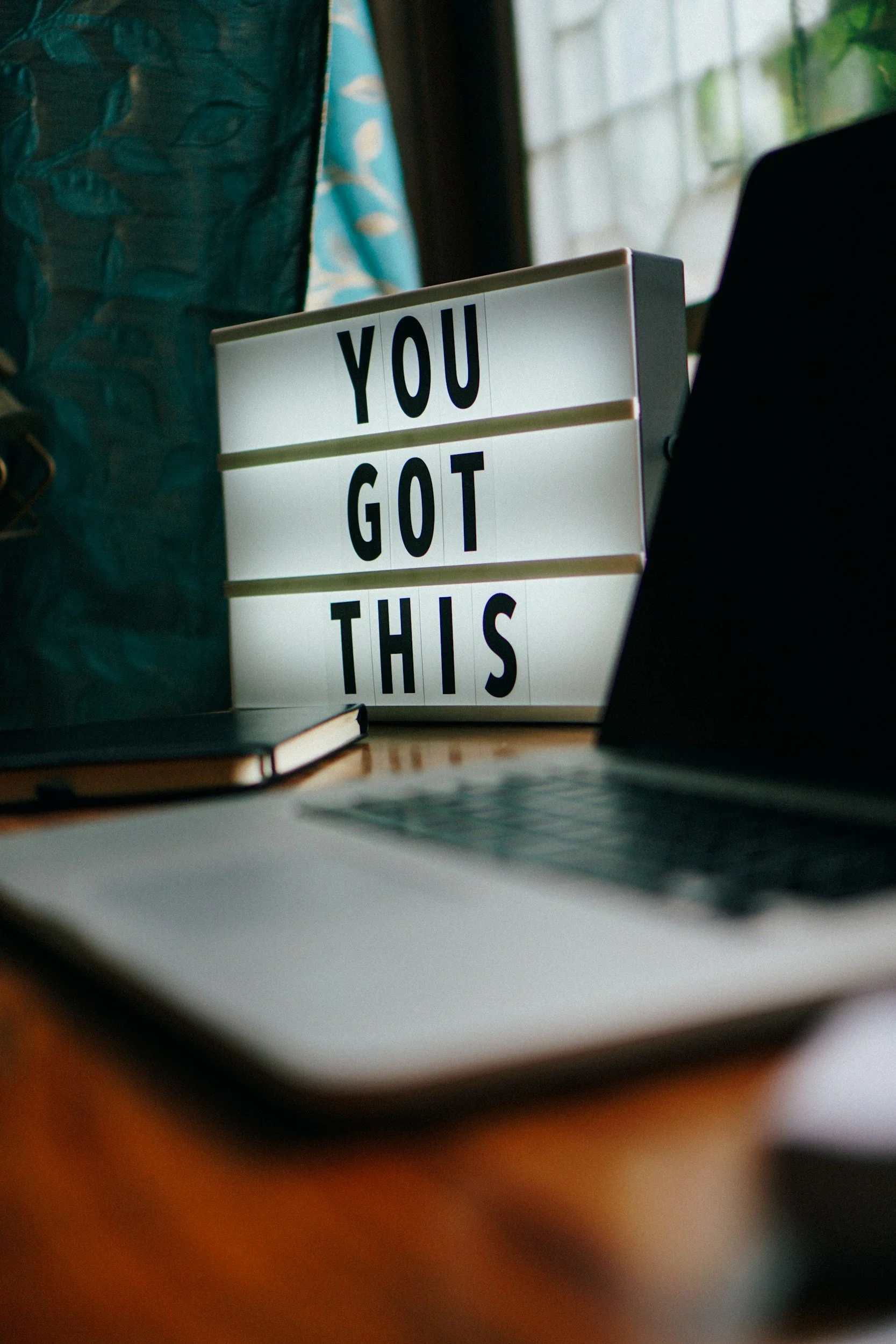 Ein beleuchtetes Schild mit der Aufschrift "YOU GOT THIS" auf einem Schreibtisch, umgeben von einem Laptop, einem Buch und einem Notizbuch