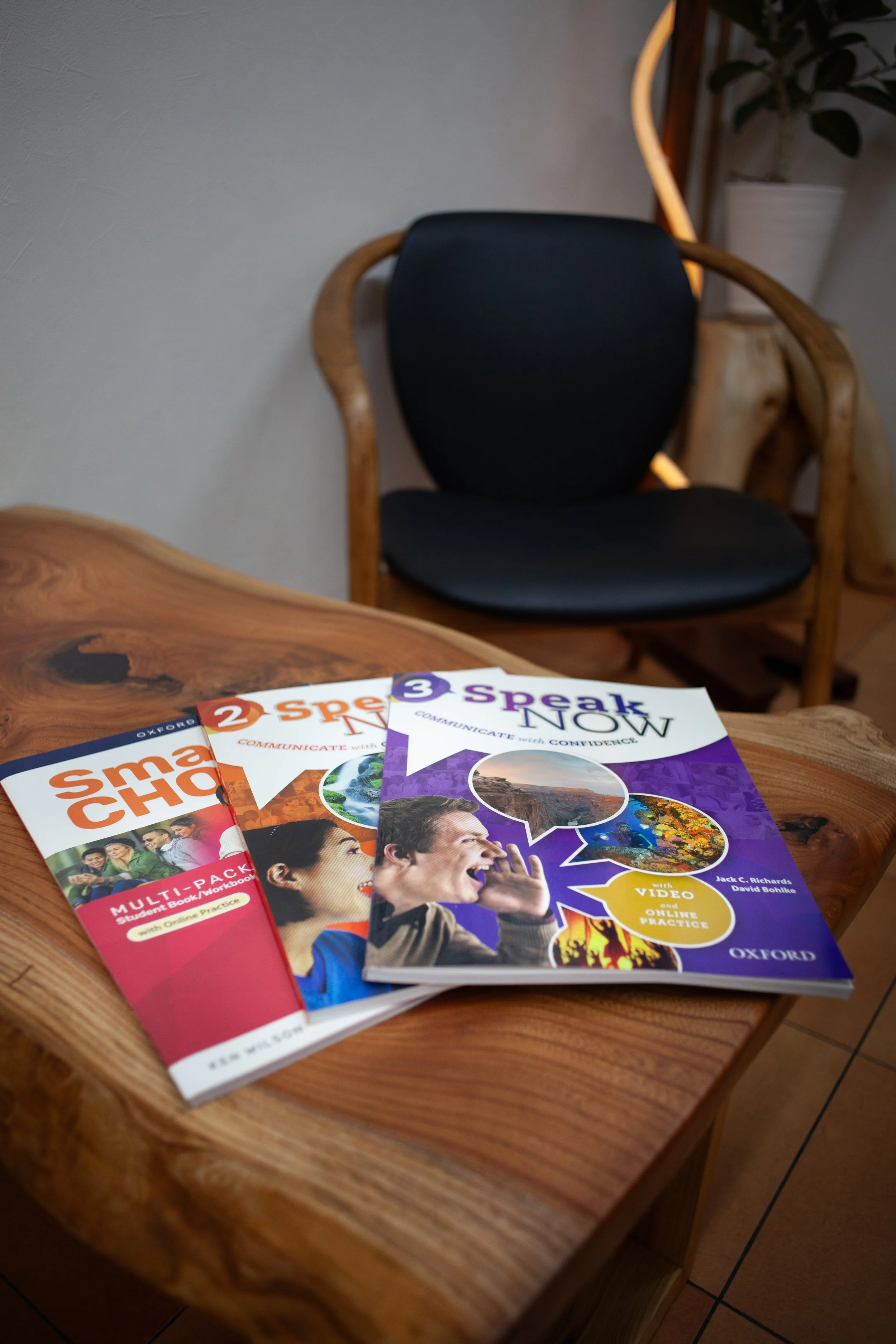 木製テーブルに並んだ3冊の英語学習テキストと、背景にある黒いアームチェアと観葉植物が映る英会話学習シーン