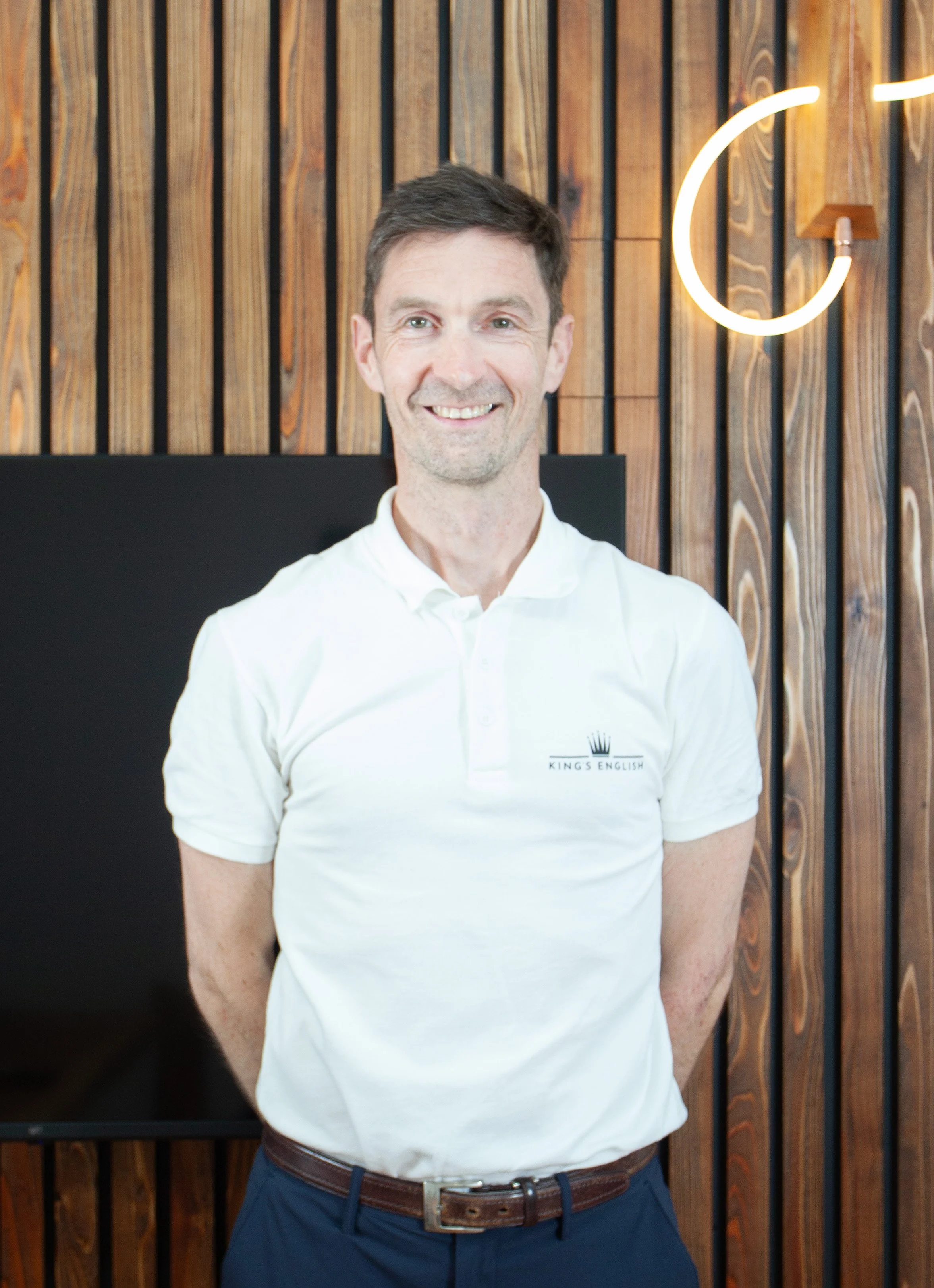 木製の壁とモダンなライトがある室内で、King's English ロゴ入り白ポロシャツを着た男性が笑顔で立つ英会話スクールのイメージ写真