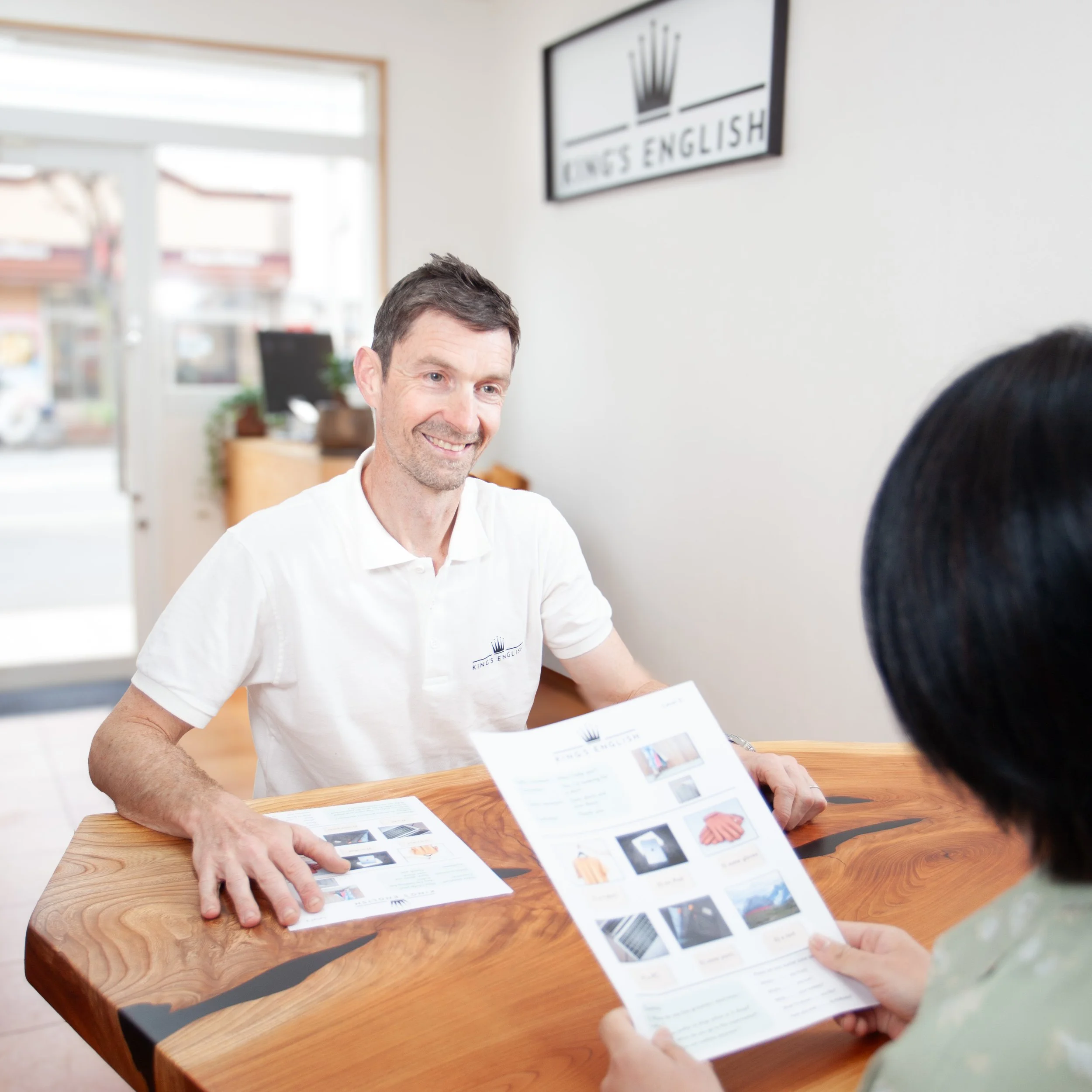 KINGS ENGLISH の看板が見える明るい英会話スクールの受付エリアで、白いロゴ入りポロシャツの男性が笑顔で女性と向かい合い、女性がパンフレットを持っている場面