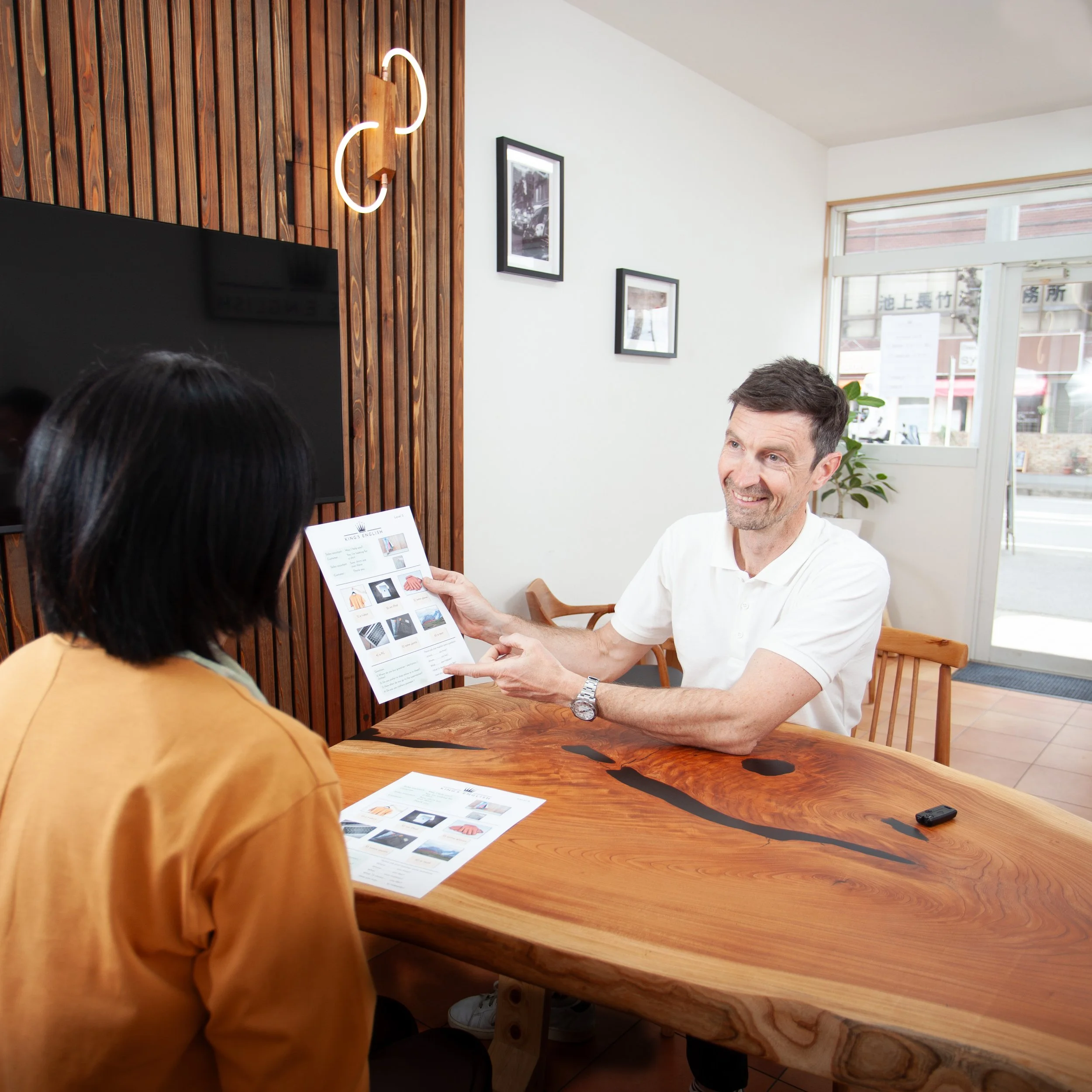 A man smiling and handing a brochure to a woman sitting at a wooden table in an English conversation school. 英会話スクールの教室で、笑顔の男性が木製テーブルに座る女性にパンフレットを手渡している場面
