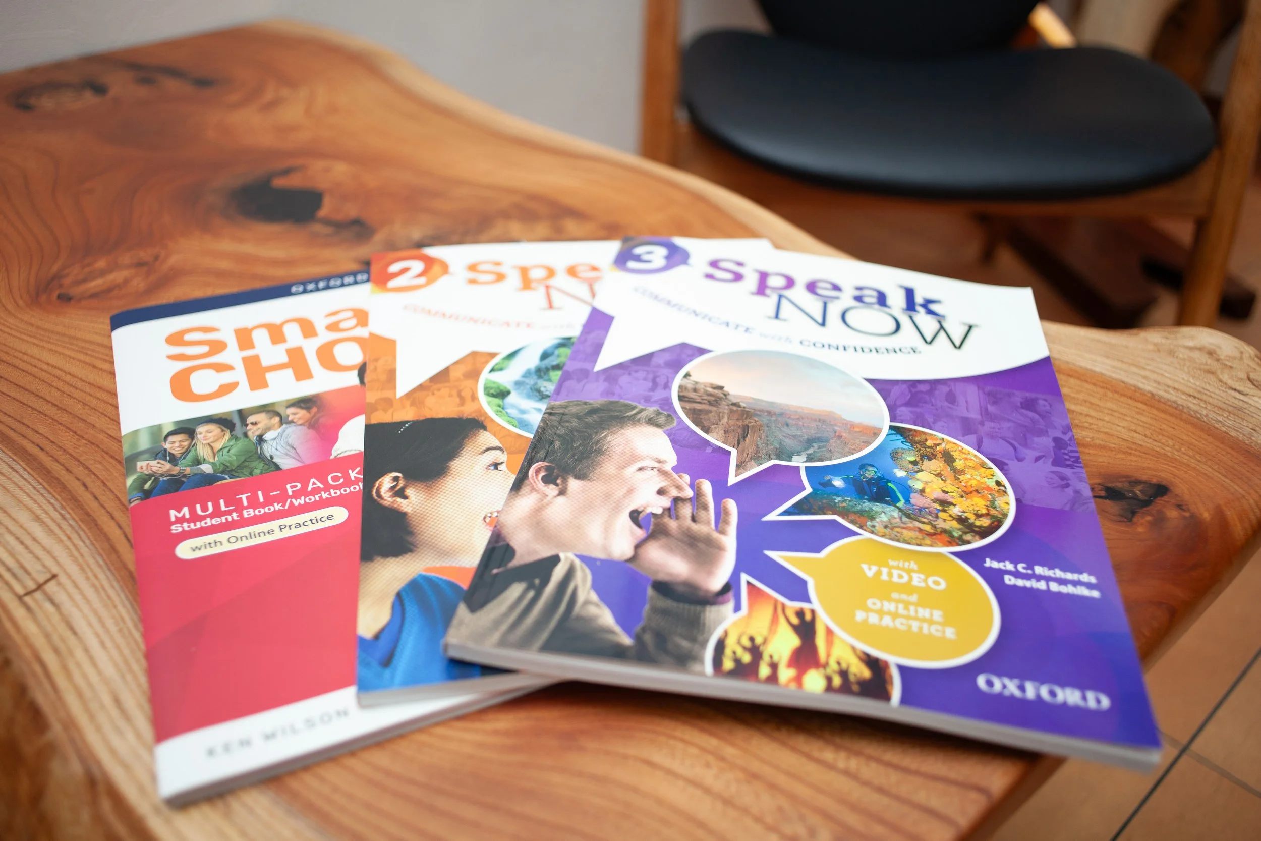 木製テーブルに置かれた Oxford Speak Now 英語学習テキスト3冊と、背景の黒い椅子が見える英会話学習シーン