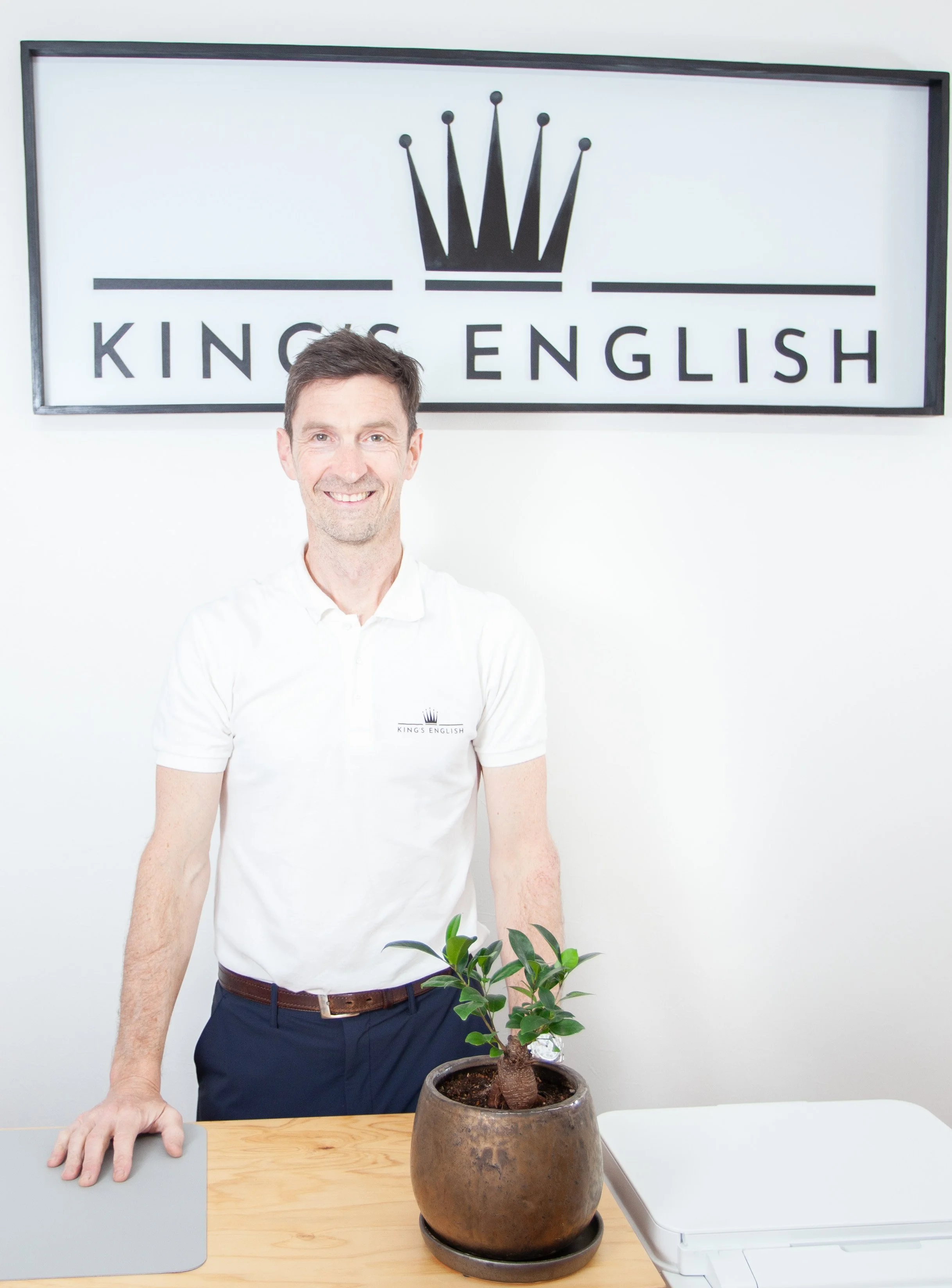 観葉植物とノートパソコン、スケールが置かれたデスクの後ろで男性が笑顔で立ち、背面には王冠ロゴ入りの “KINGS ENGLISH” 看板が掲示された英会話スクールのイメージ写真