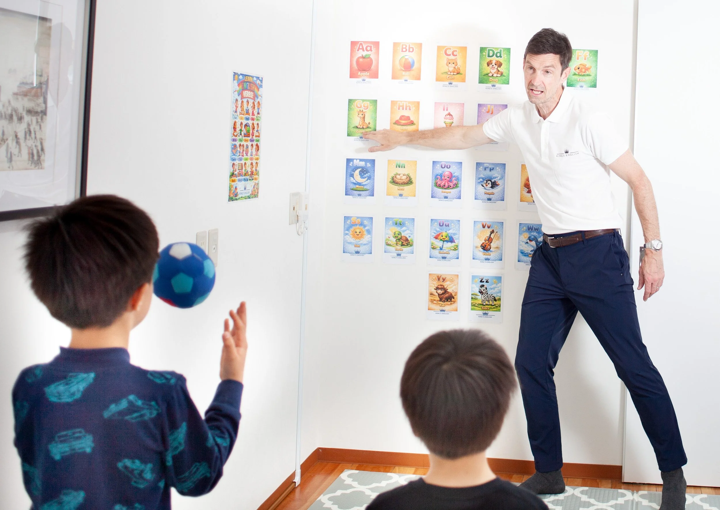 カラフルなアルファベットポスターが並ぶ教室で、講師が子どもたちにボールを使って英語を教えている子ども英会話レッスンの場面