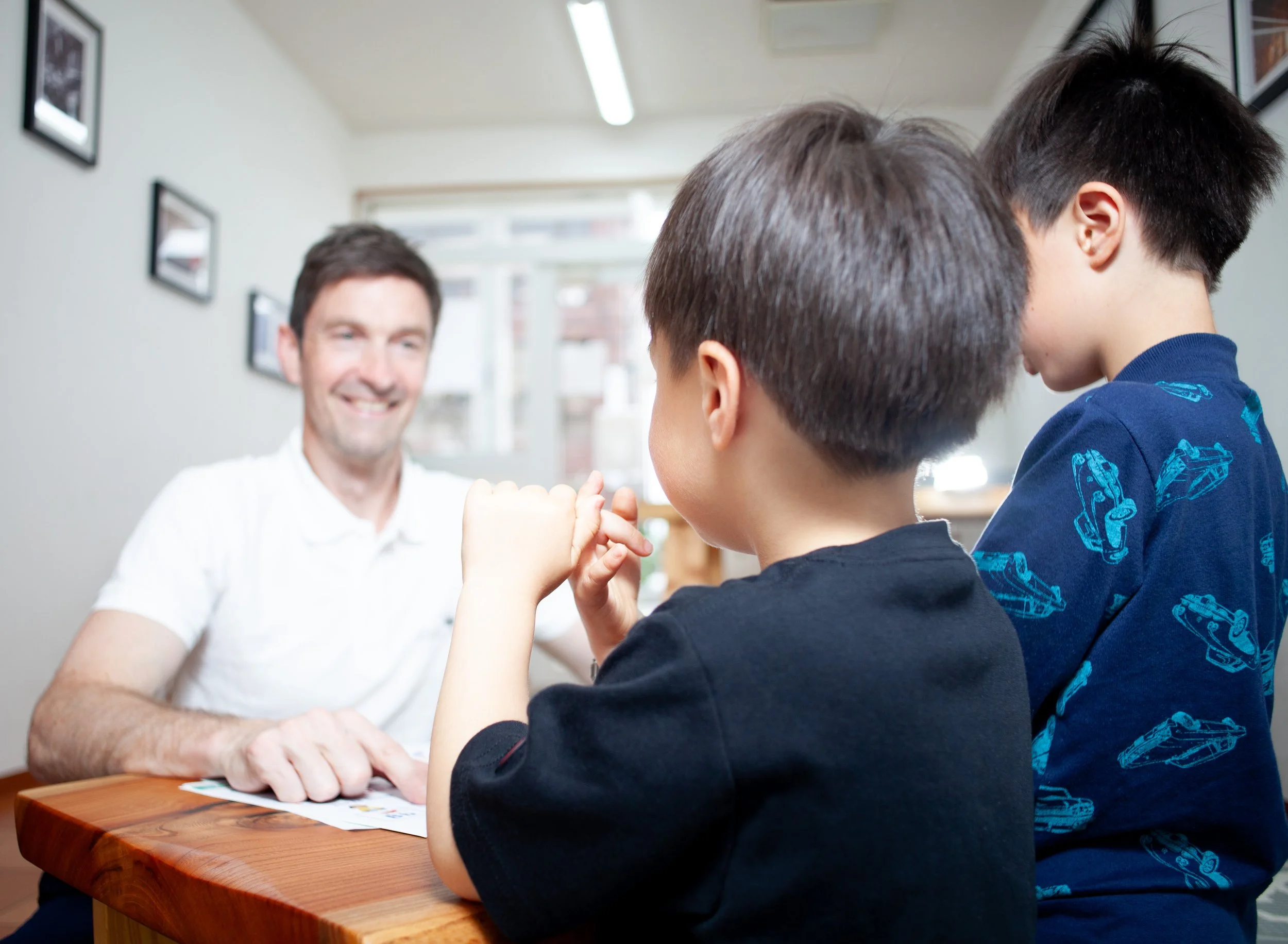 明るい教室で、男性が2人の少年と木製テーブルを囲んで会話やアクティビティを行い、壁には額入り写真が並ぶ子ども向け英会話シーン