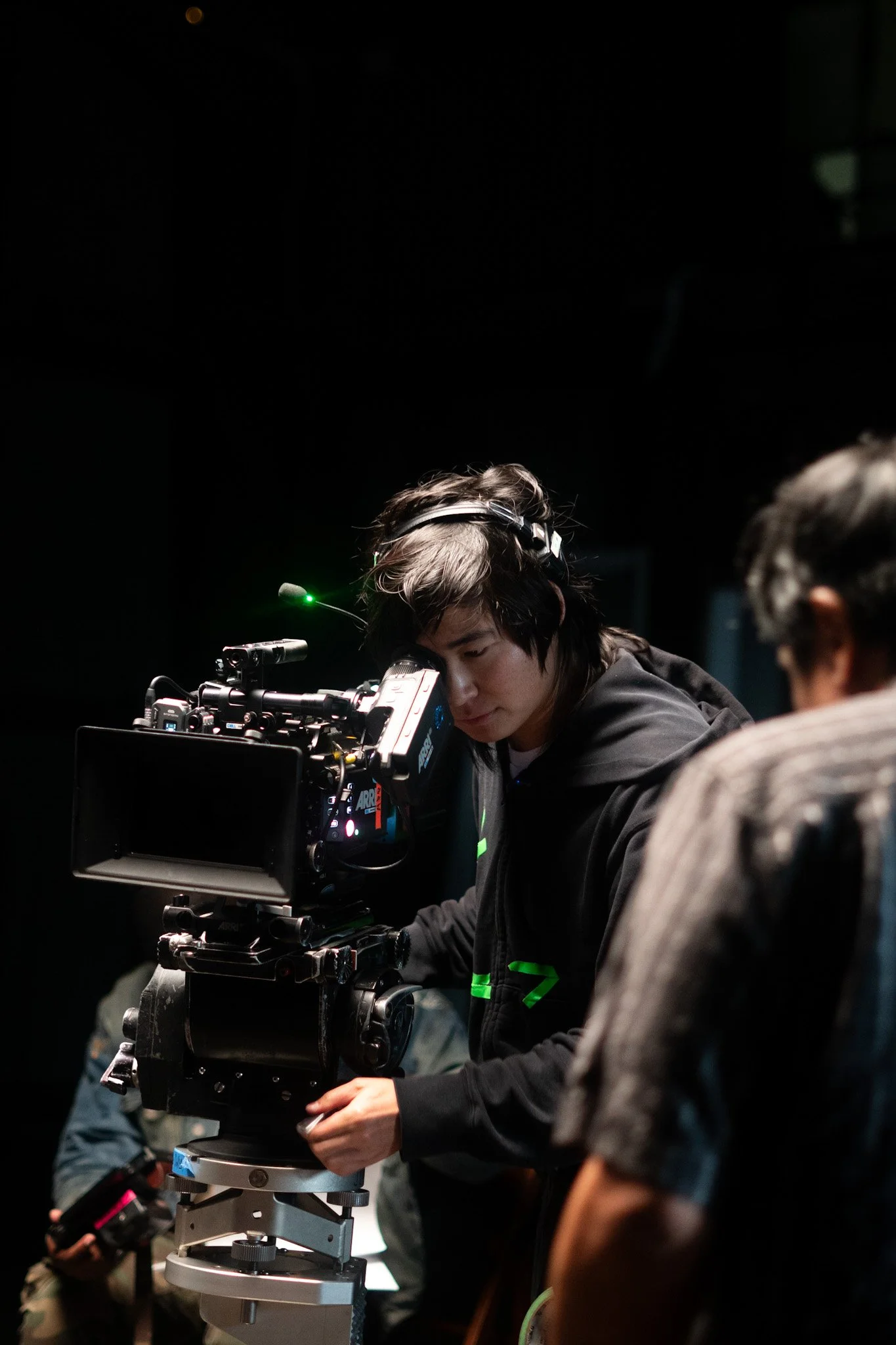 A person with dark hair wearing a black jacket is looking through a camera mounted on a tripod, in a dark setting with other people around.