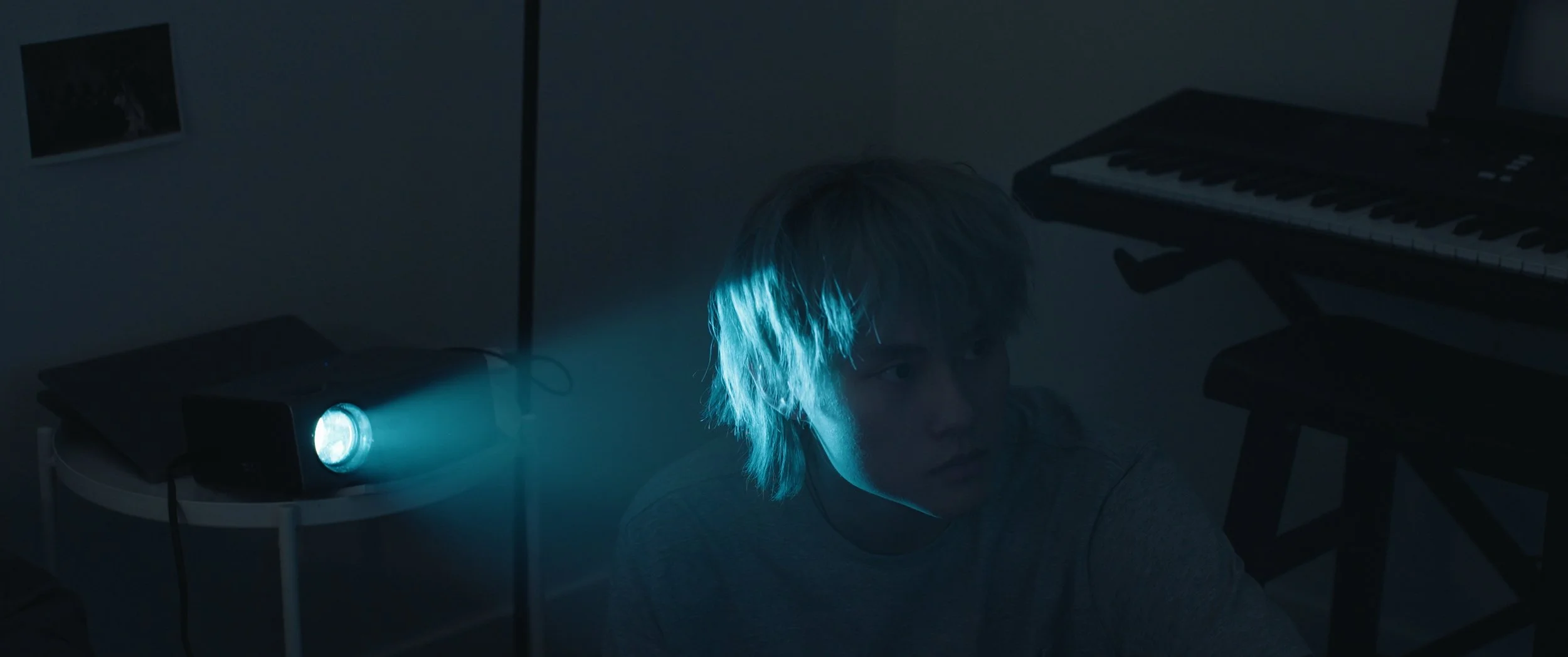 A person with light-colored hair looking over their shoulder in a dimly lit room, illuminated by a blue spotlight on a small table, with a keyboard instrument in the background.