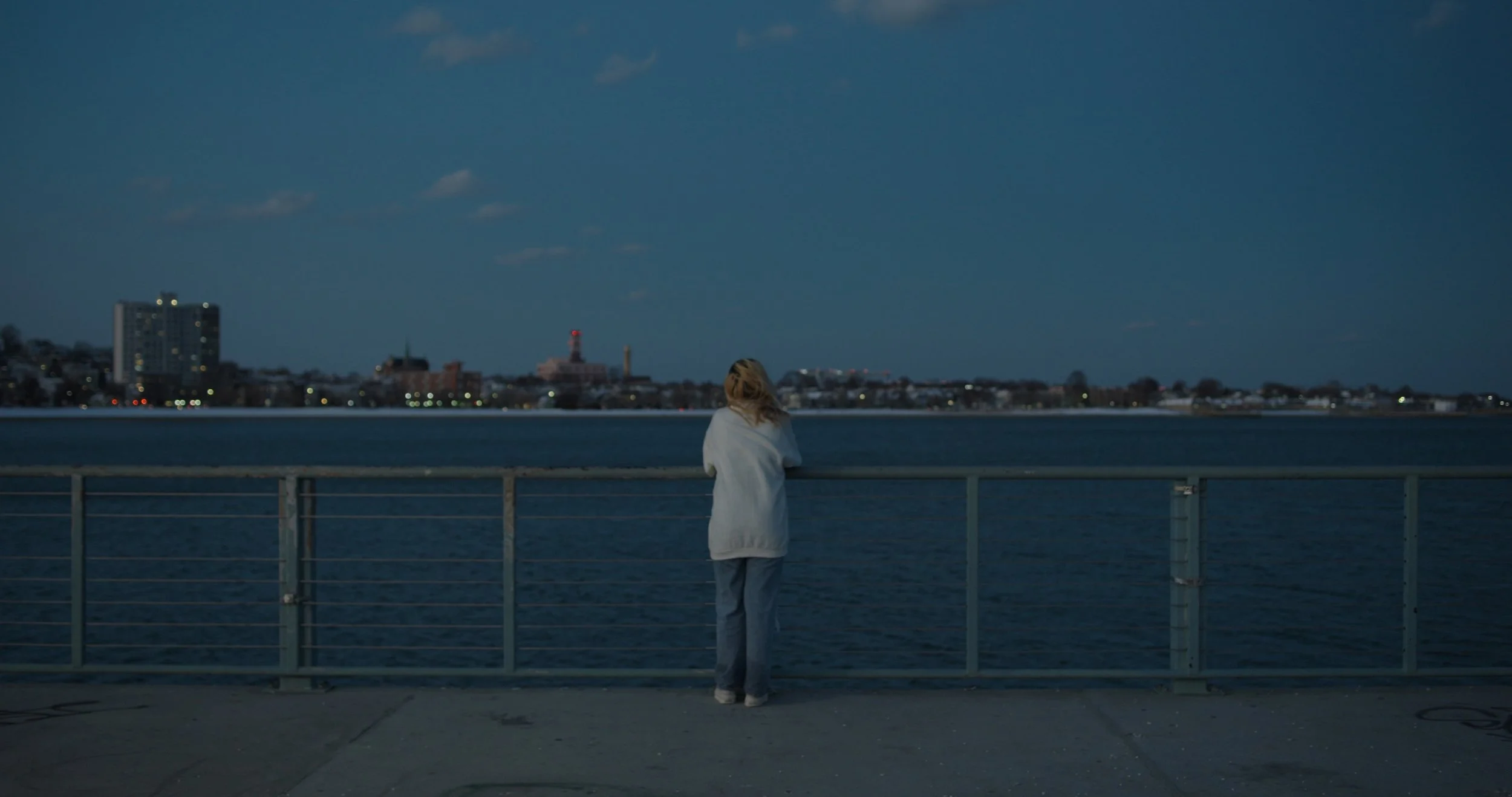 A woman with blonde hair, dressed in a light-colored coat and pants, stands alone on a waterfront deck at dusk, gazing across the water toward a distant city skyline with buildings and a lighthouse.