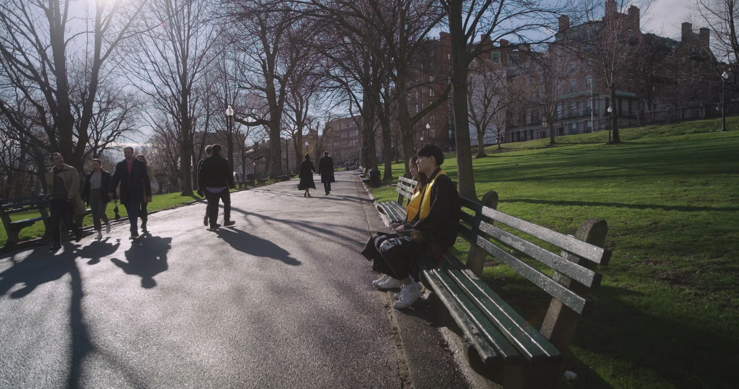 Park scene with people walking and sitting on benches, leafless trees, and buildings in the background, on a sunny day.